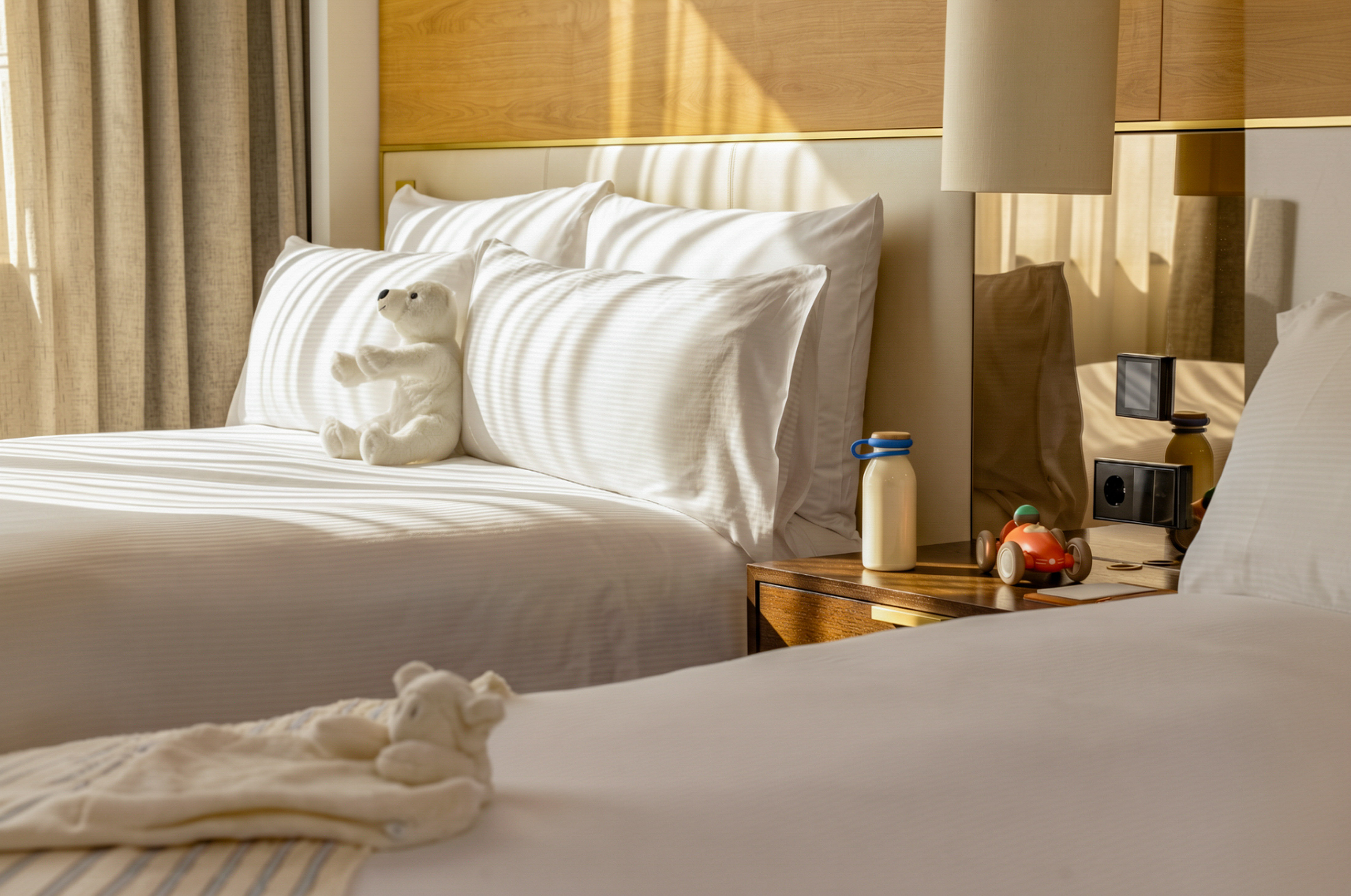 Family hotel room with twin beds, white bedding, teddy bear, and toy car.