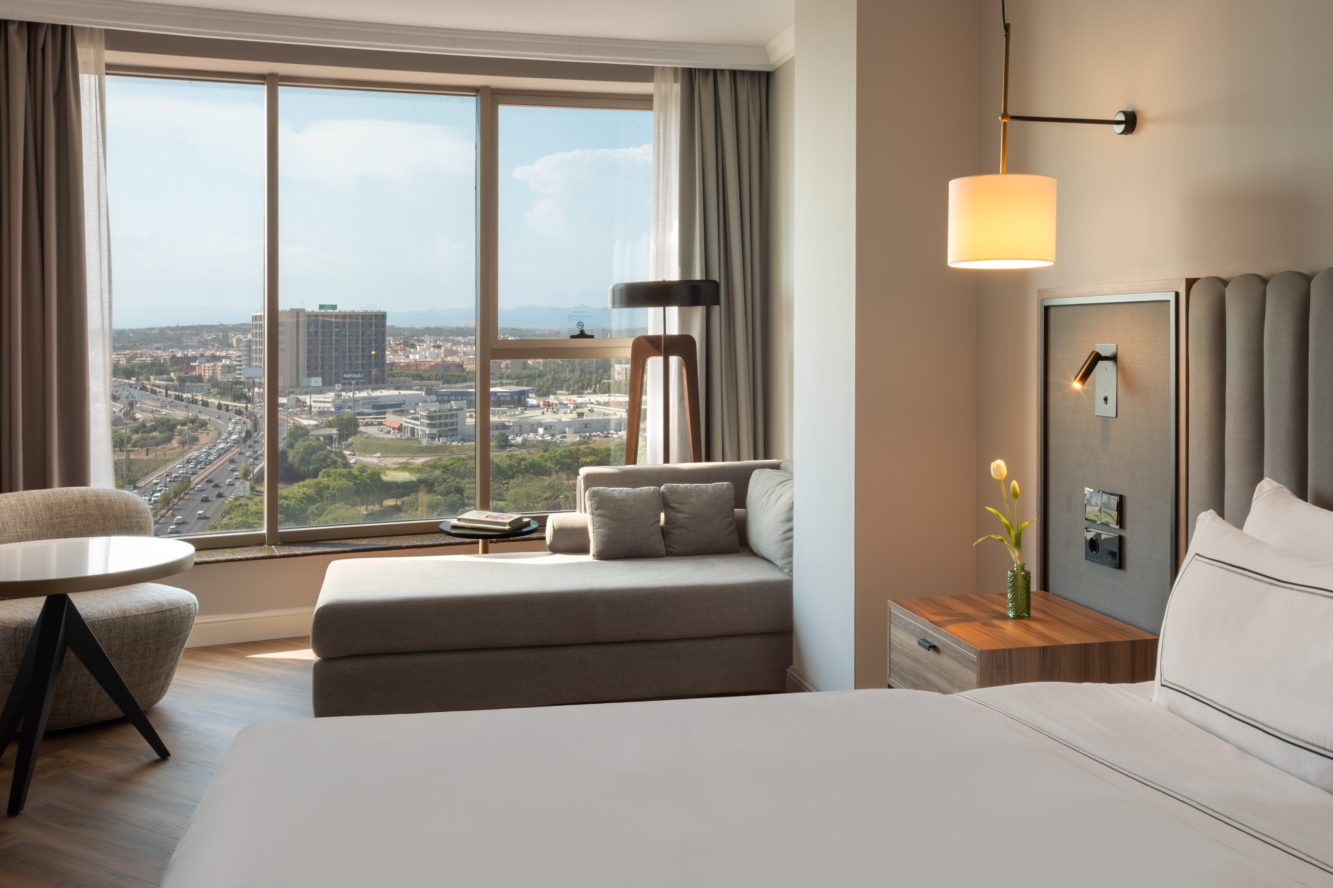 Suite de hotel moderna con vista panorámica a la ciudad, cama y chaise longue.