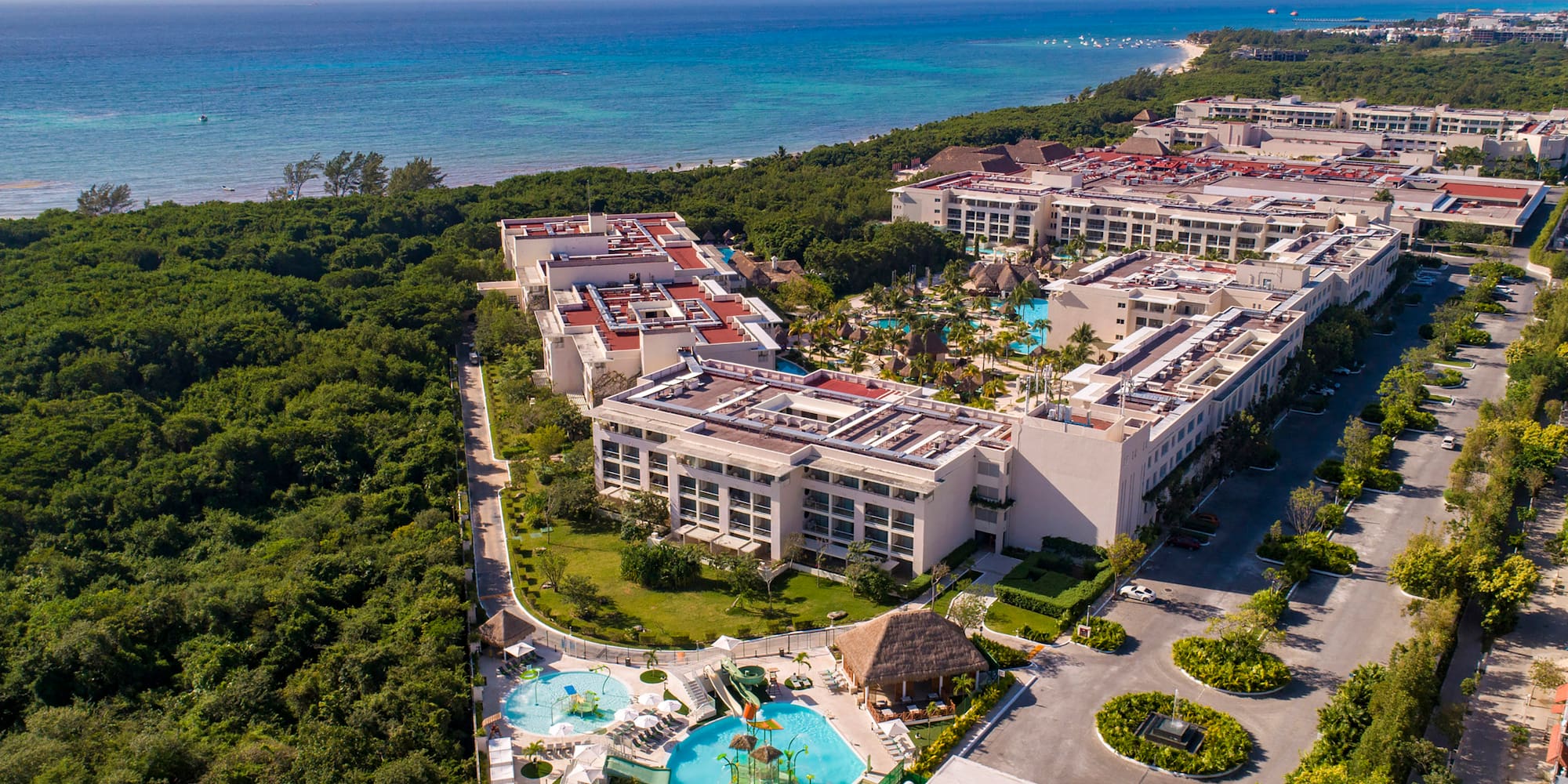 Beachfront resort aerial: Pools, water park, lush forest, blue ocean.