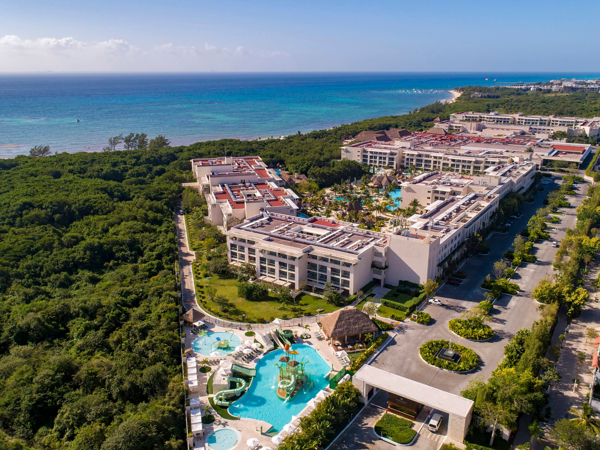 Beachfront resort aerial: Pools, water park, lush forest, blue ocean.