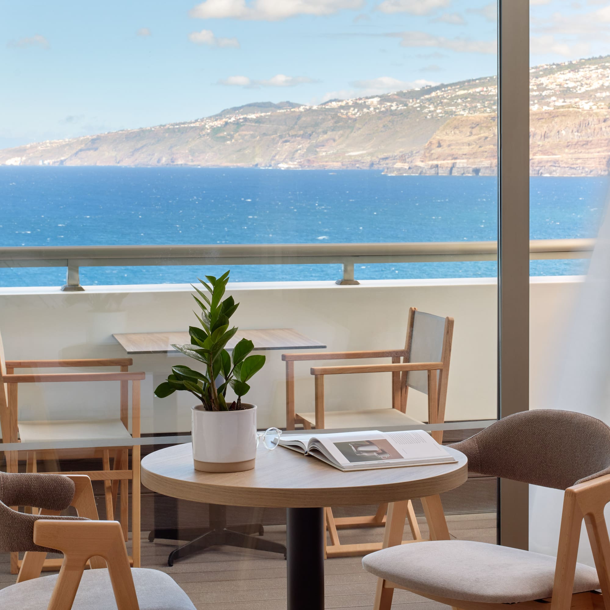 a table and chairs with a book on it by a window overlooking the ocean