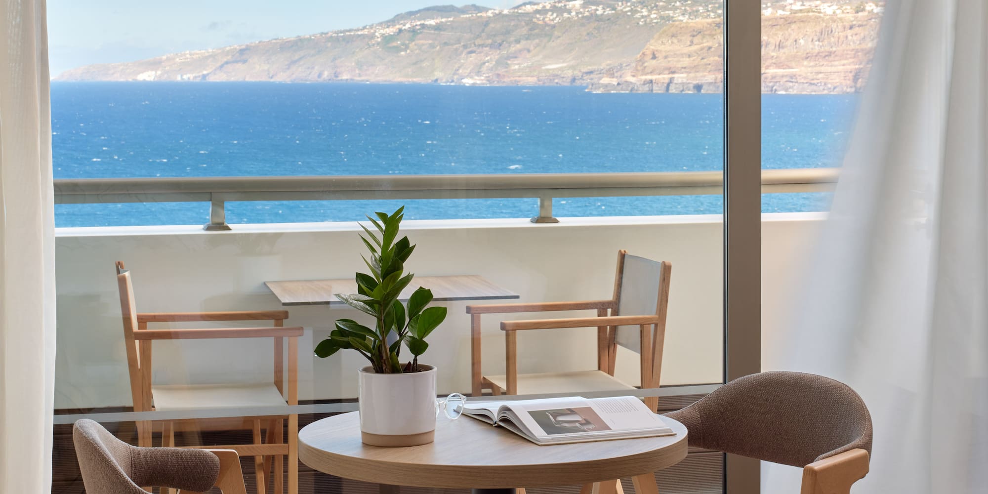 a table and chairs with a book on it by a window overlooking the ocean