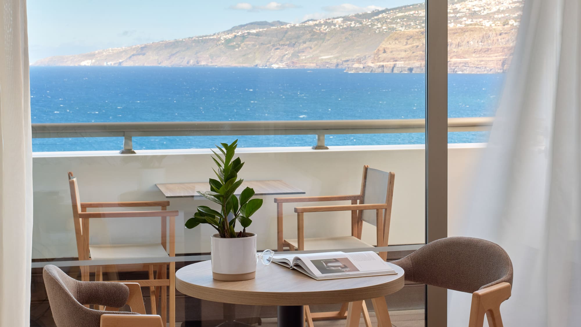 a table and chairs with a book on it by a window overlooking the ocean