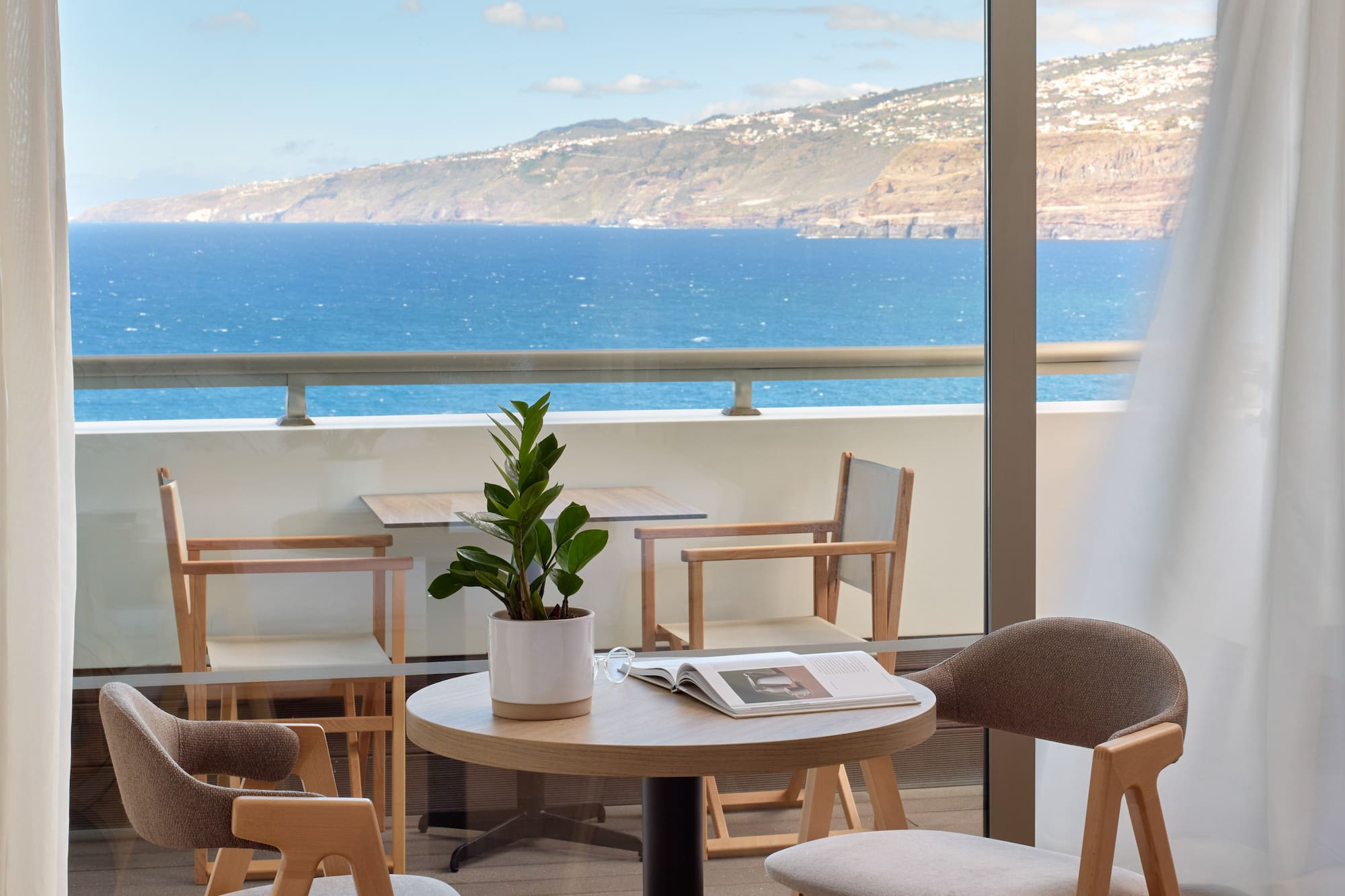 a table and chairs with a book on it by a window overlooking the ocean