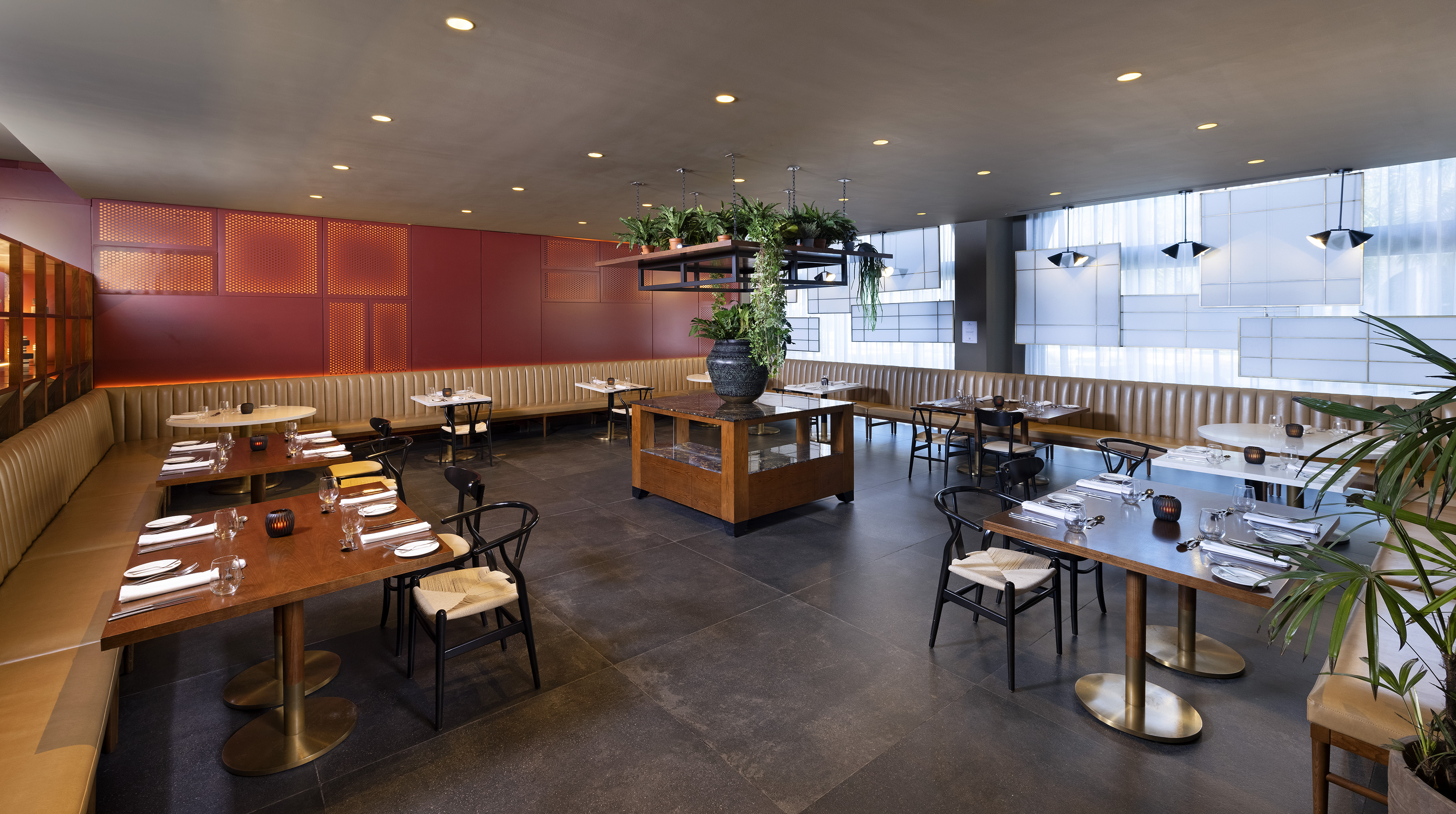 Modern restaurant with red accent wall, leather banquettes, wooden tables, and plants.