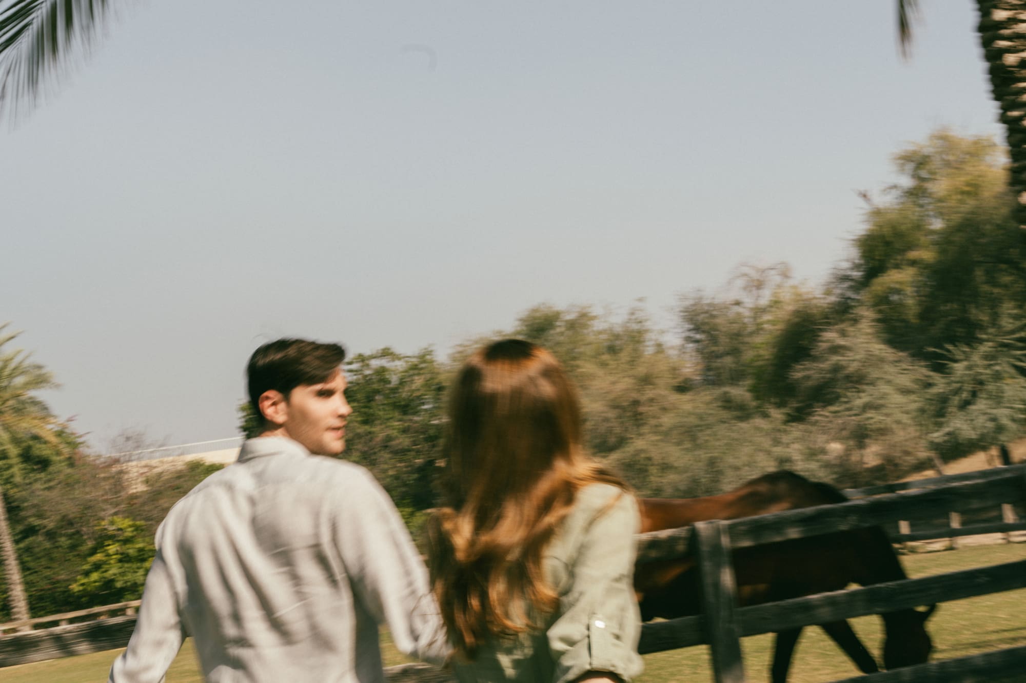 Couple strolling by horse fence and palm trees at a sunny ranch.