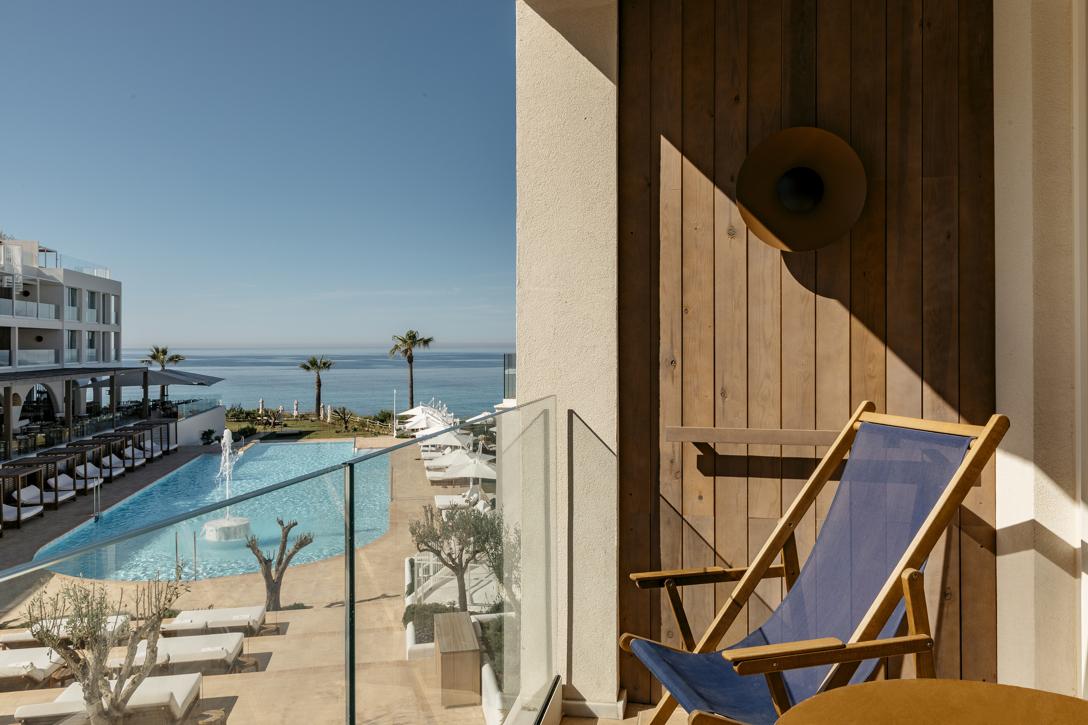 Resort balcony with ocean view, swimming pool, palm trees, and deck chair.