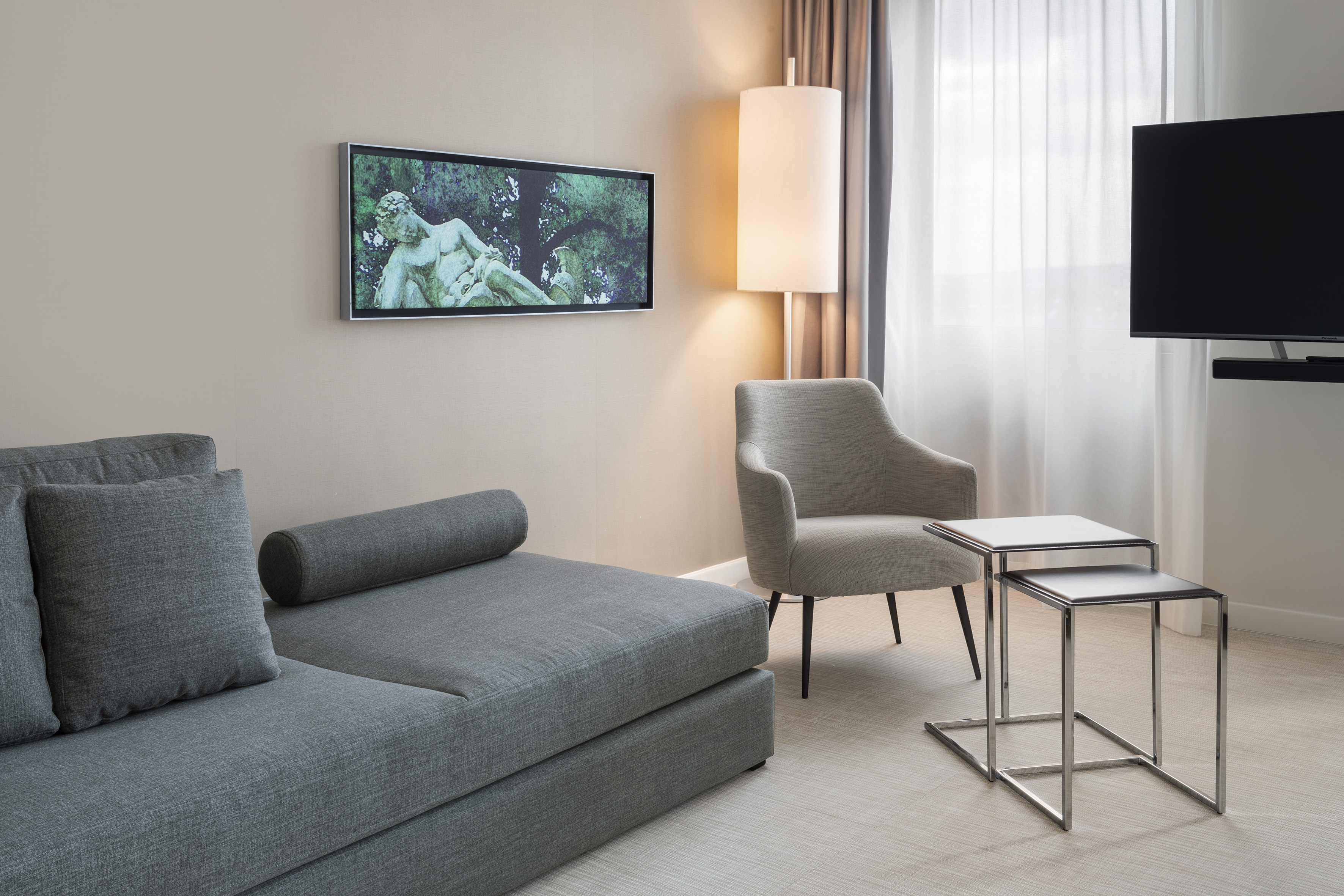 Modern hotel living area with grey sectional sofa, wall-mounted art TV, and armchair.