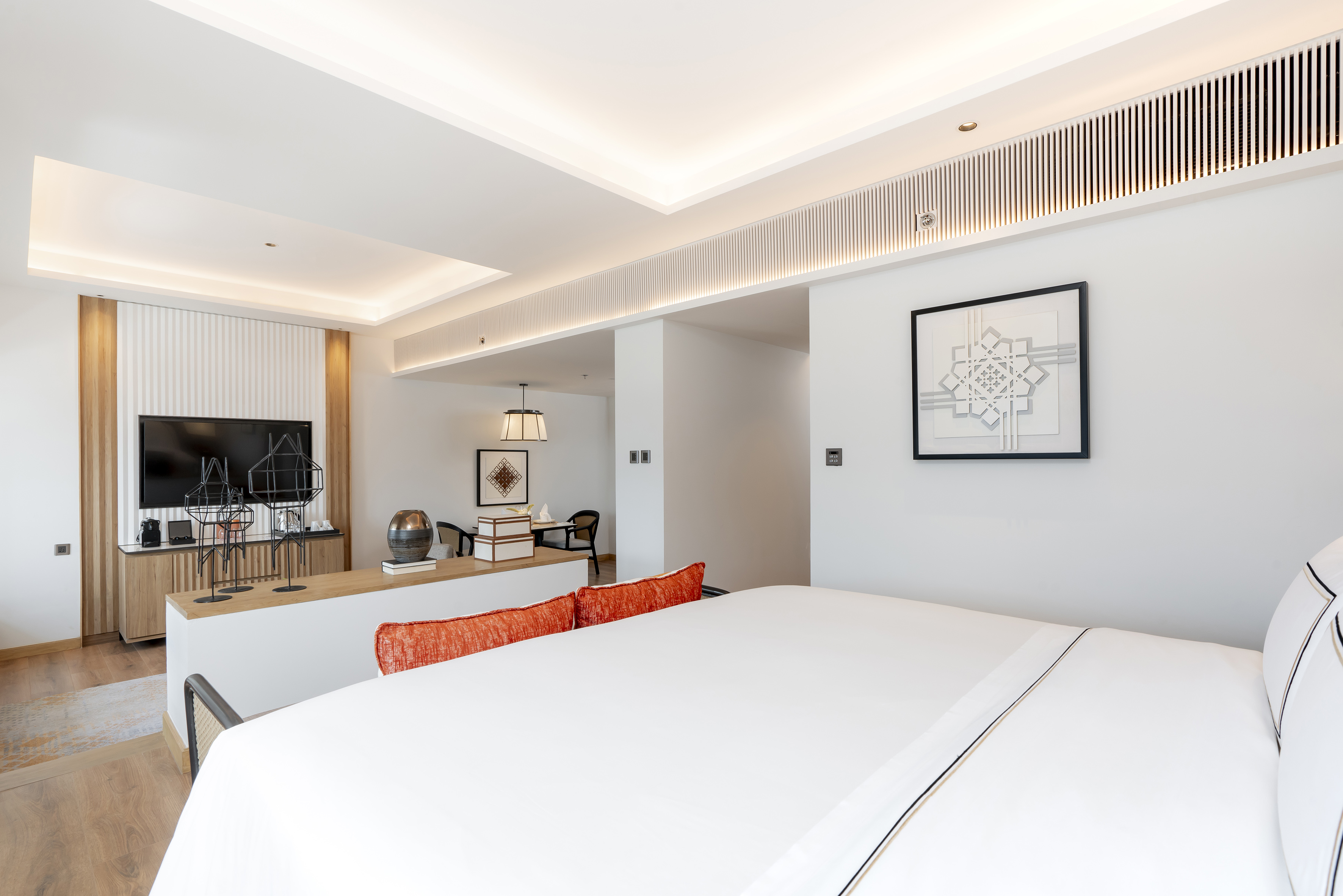 Modern hotel suite with white bed, orange cushions, TV, and open layout.