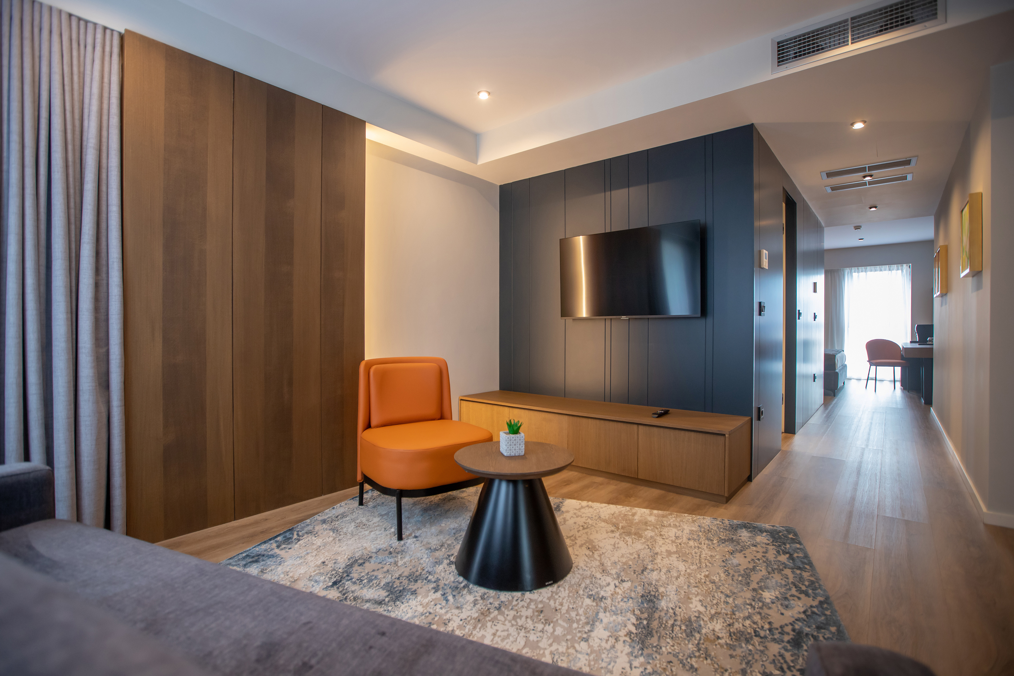 Modern hotel suite with orange accent chair, wall TV, and wood paneling.