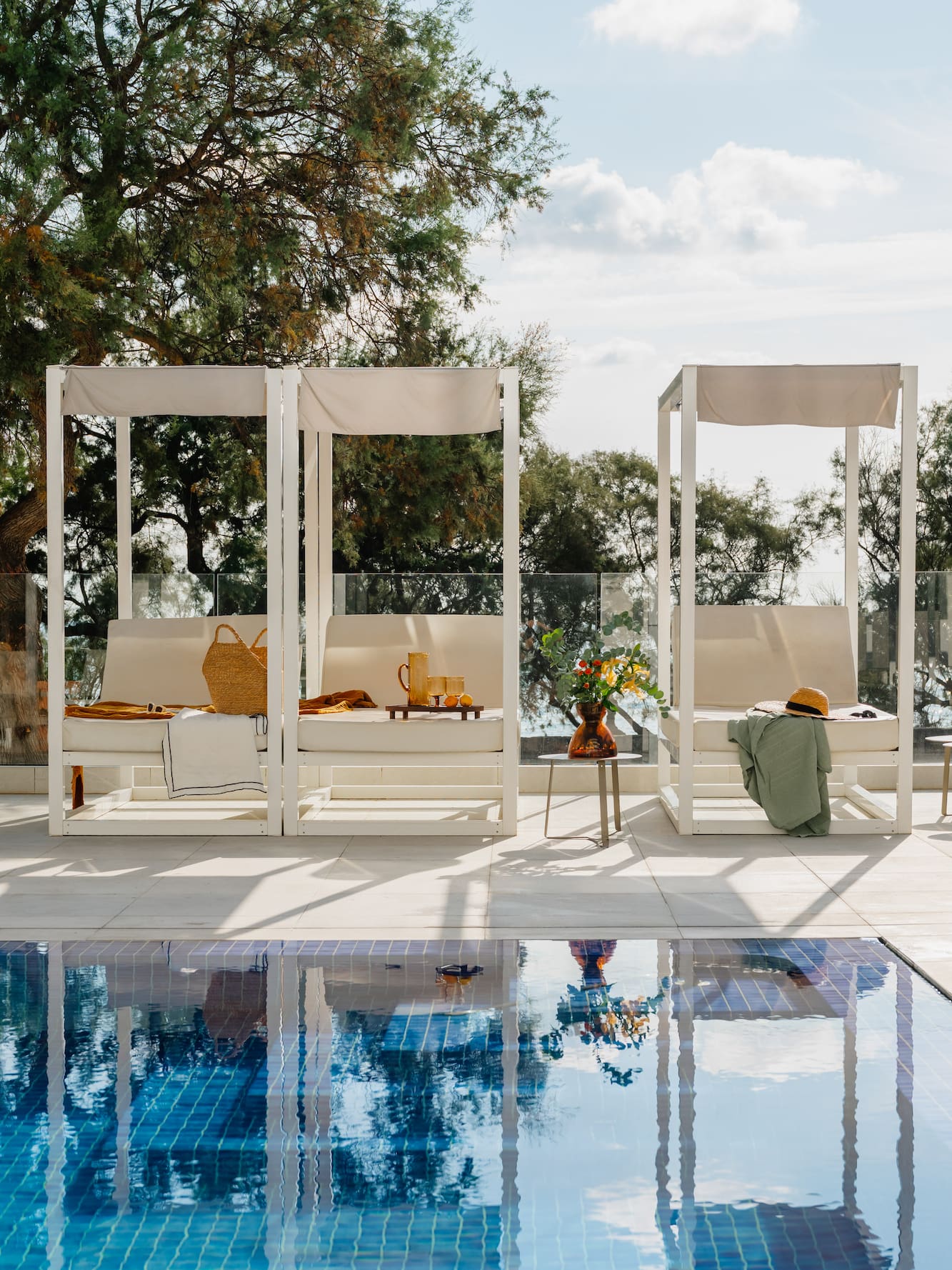 White cabanas with daybeds, straw bag, drinks by a reflective blue pool.