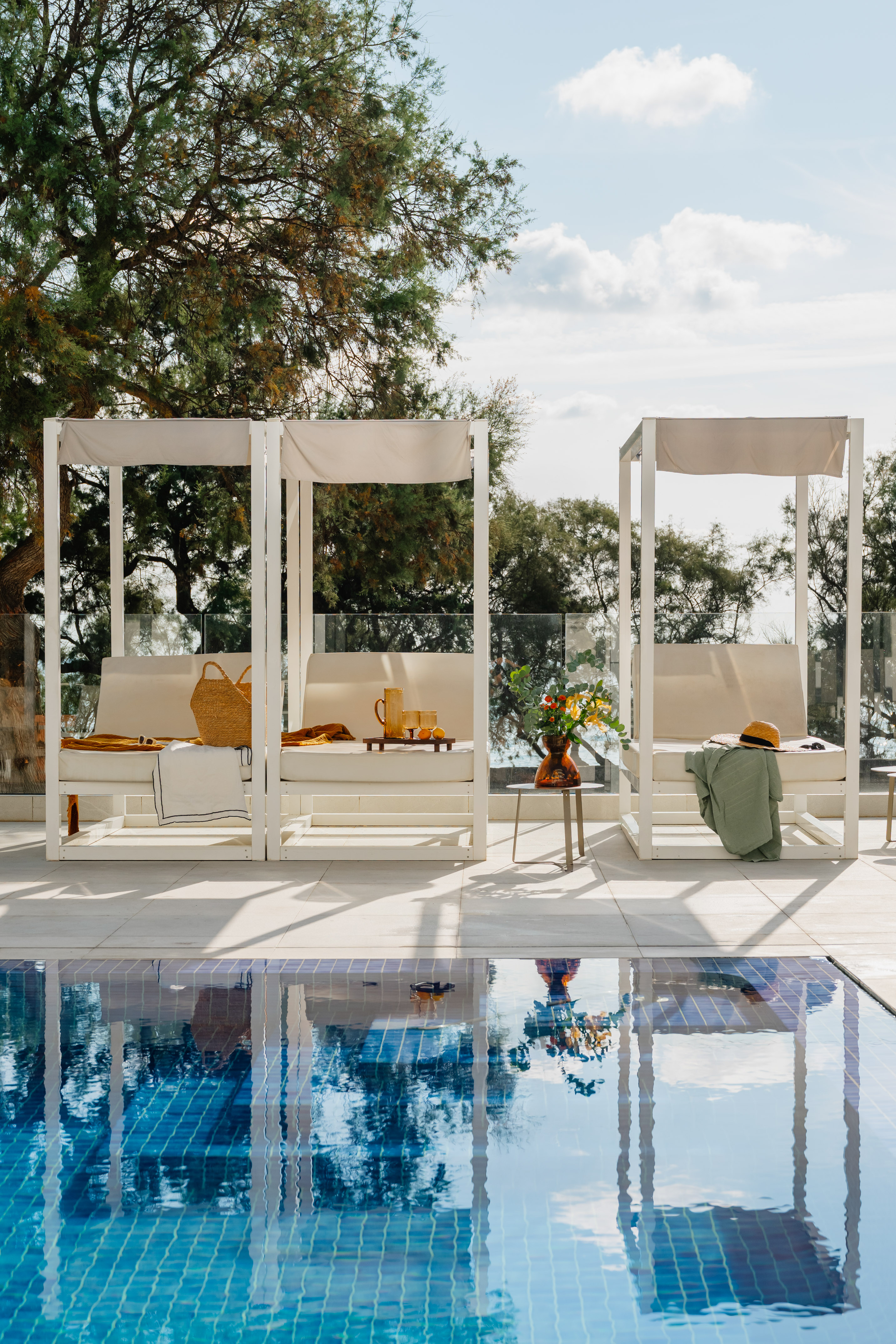 White cabanas with daybeds, straw bag, drinks by a reflective blue pool.