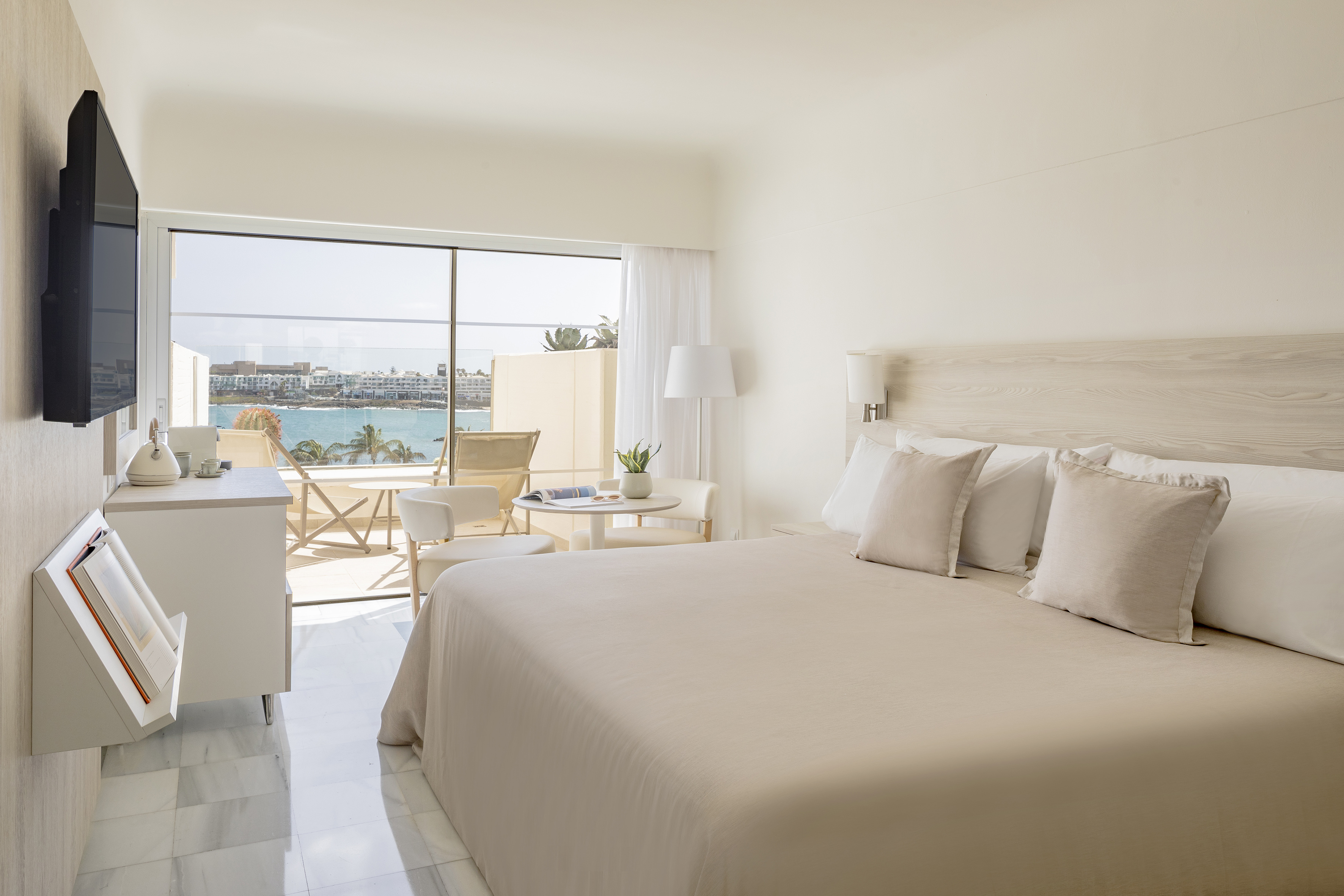 Modern hotel room with large bed, sea view balcony, and bright coastal decor.