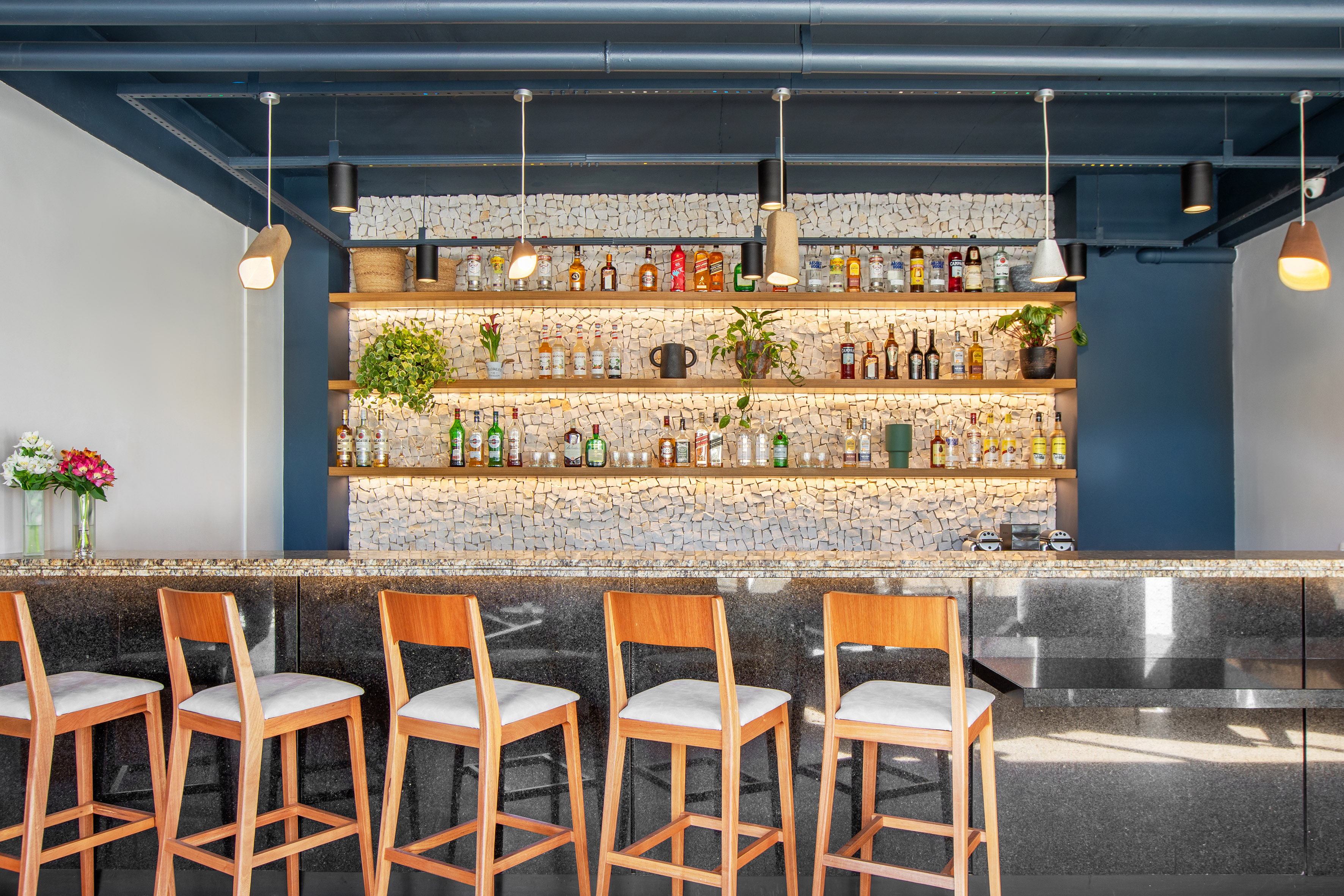 Modern bar with dark granite counter, wooden stools, backlit liquor, and mosaic wall.