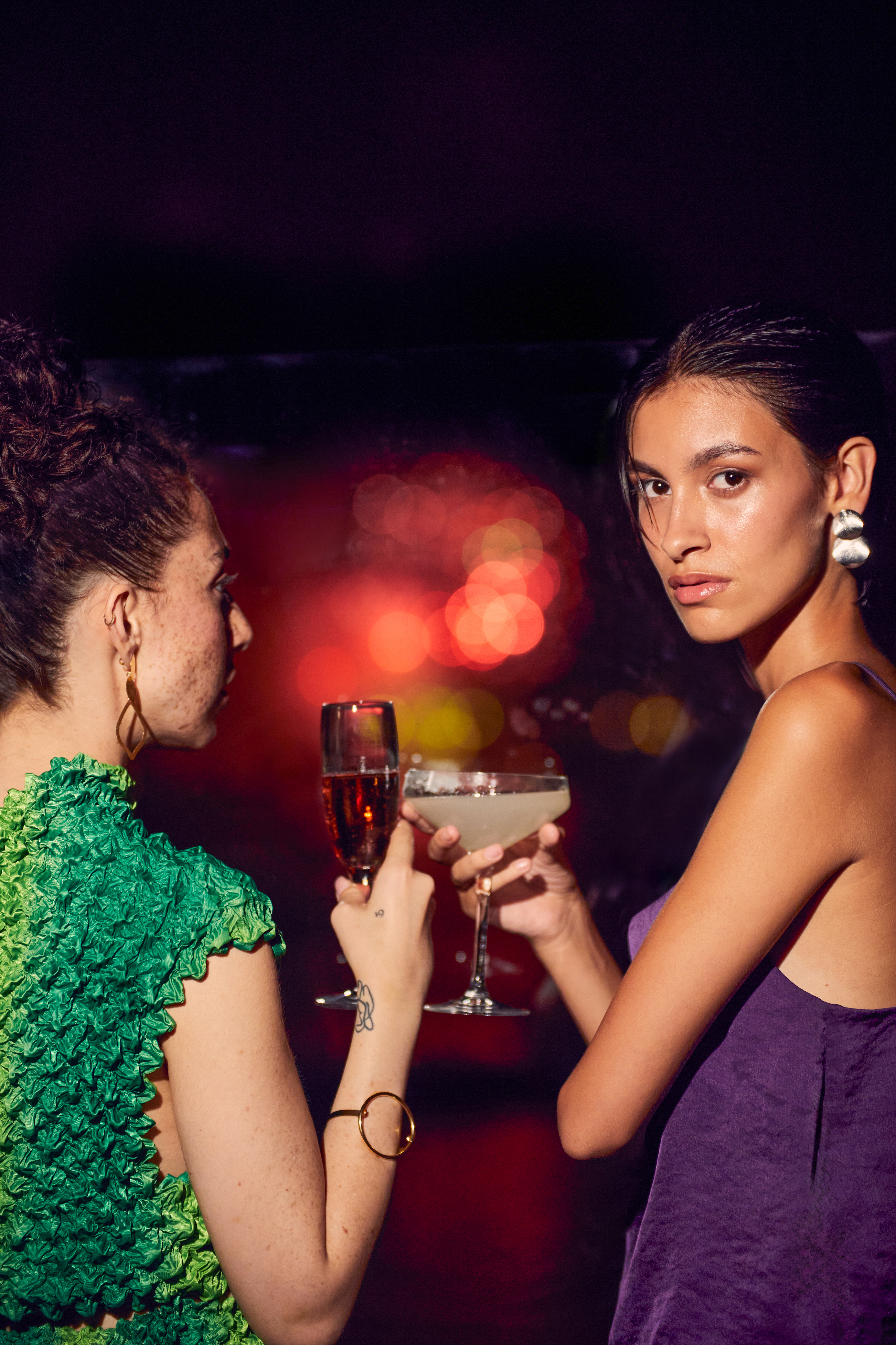 Two women at a bar toasting cocktails, vibrant bokeh lights in background.