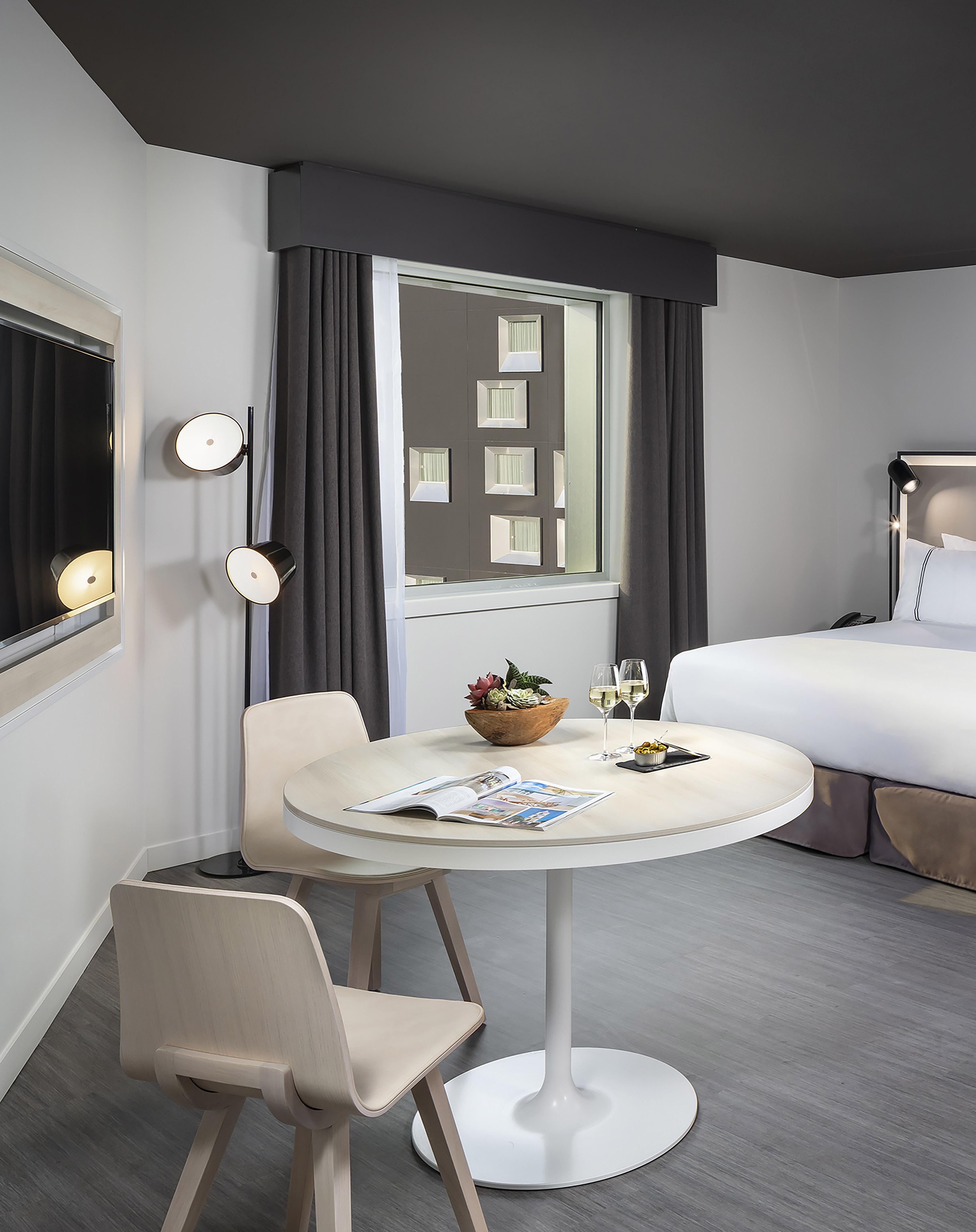 Modern hotel room with bed, round table with wine, two chairs, and window view.