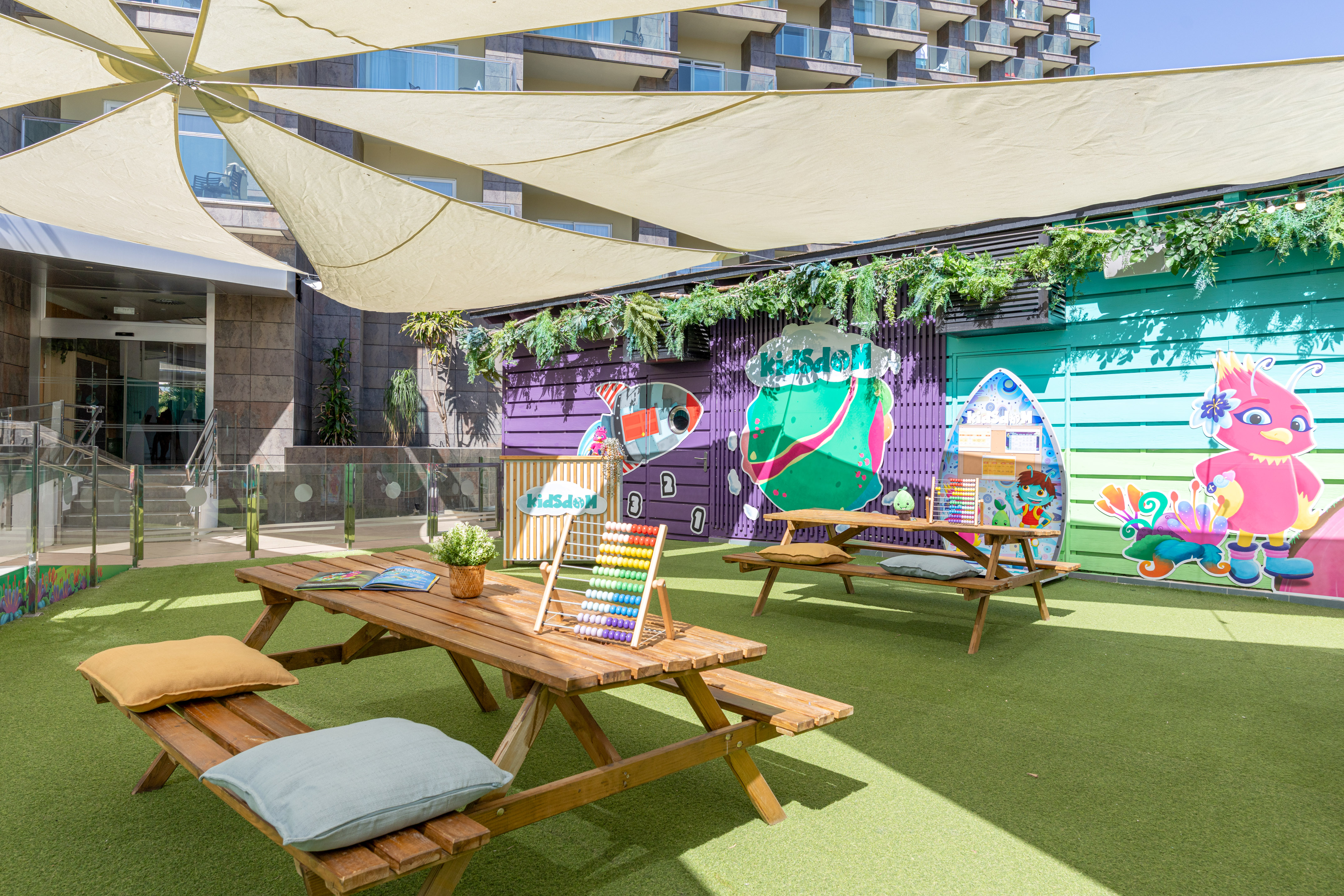 Shaded outdoor kids' club with colorful murals, picnic tables, and green turf.