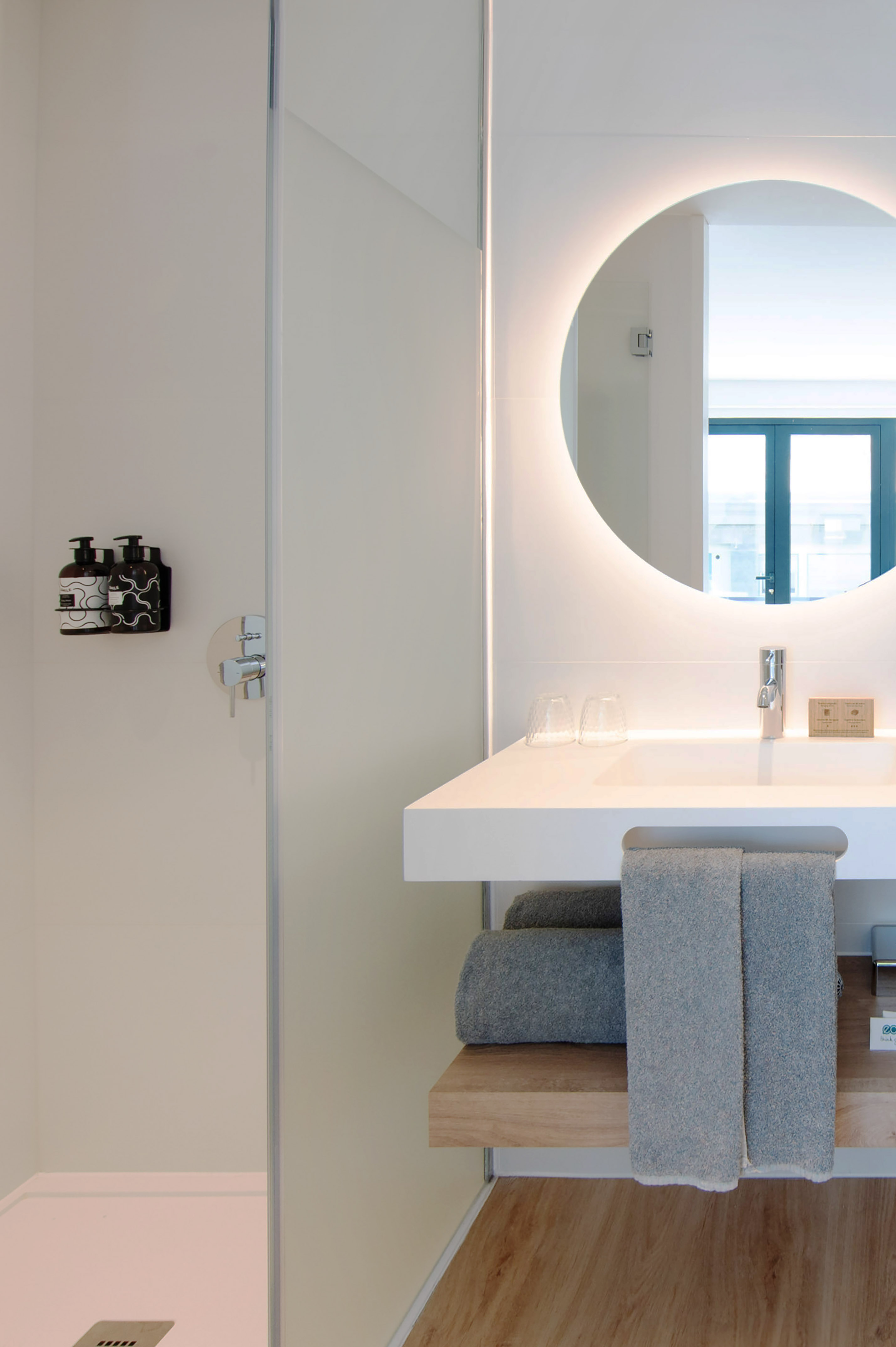 Modern hotel bathroom with illuminated round mirror, white vanity, and shower.