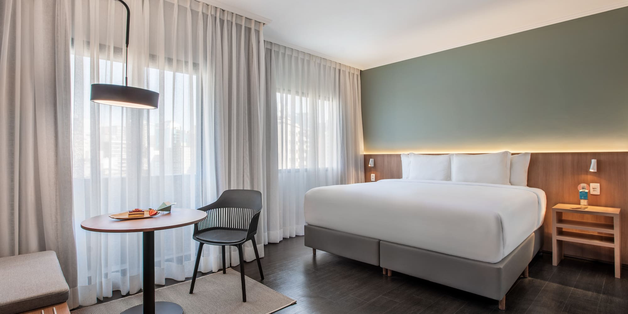 Modern hotel room with large bed, green wall, sheer curtains, and small table.
