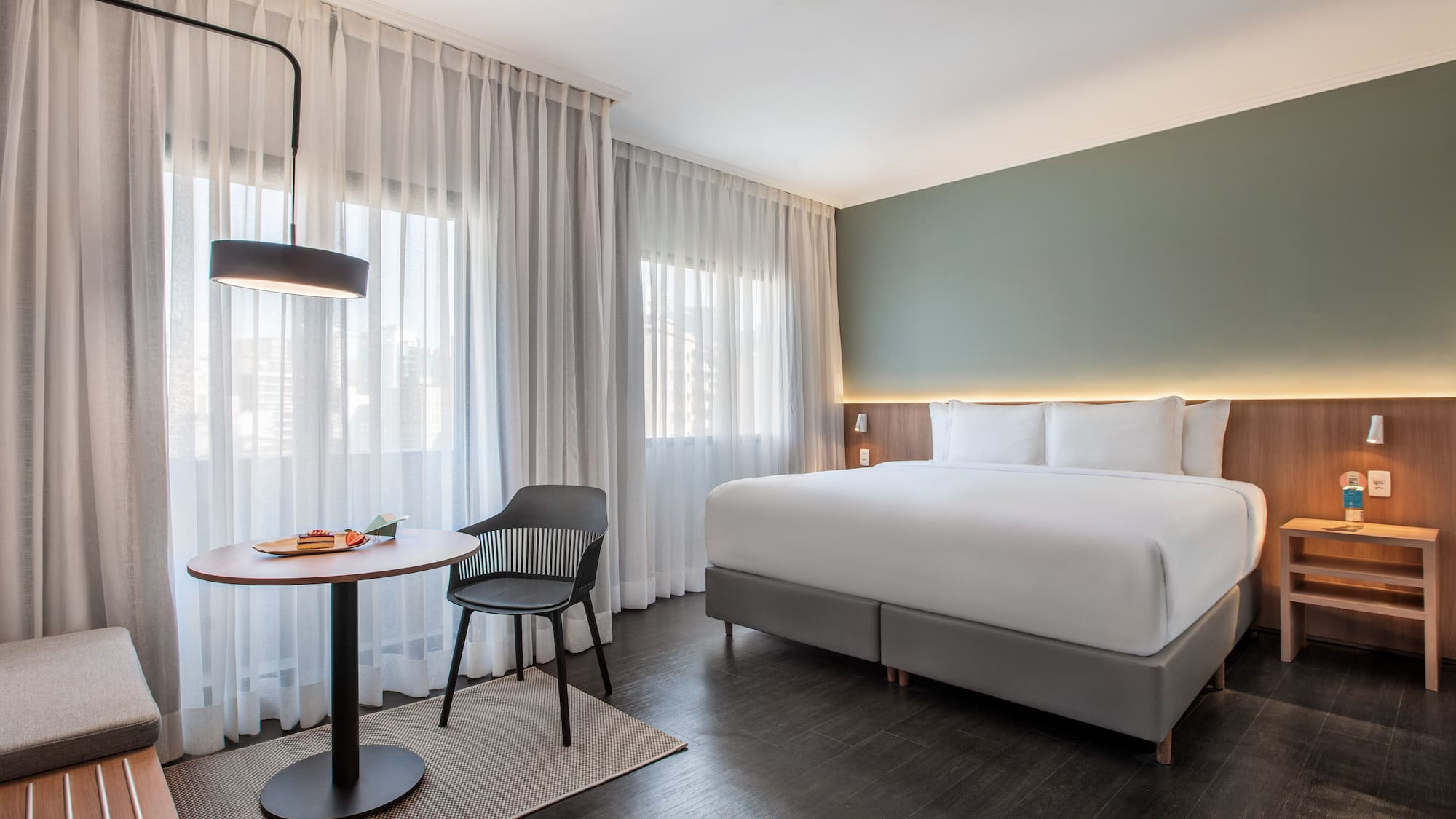 Modern hotel room with large bed, green wall, sheer curtains, and small table.