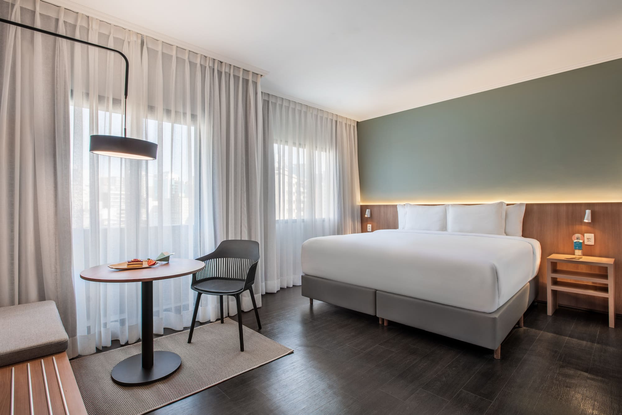 Modern hotel room with large bed, green wall, sheer curtains, and small table.