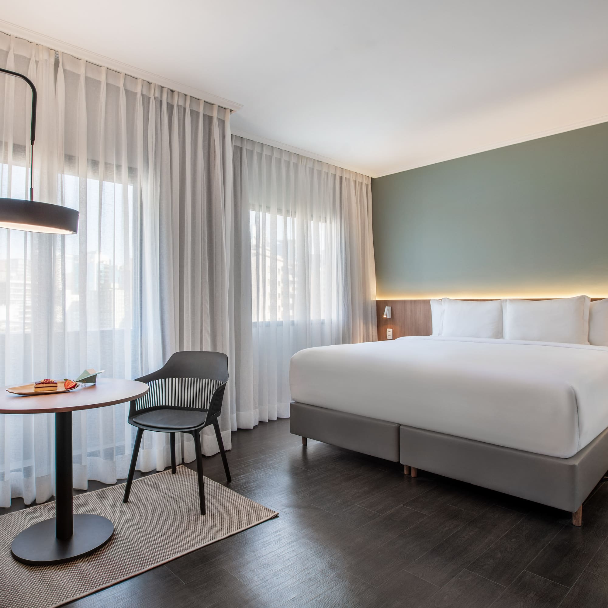 Modern hotel room with large bed, green wall, sheer curtains, and small table.