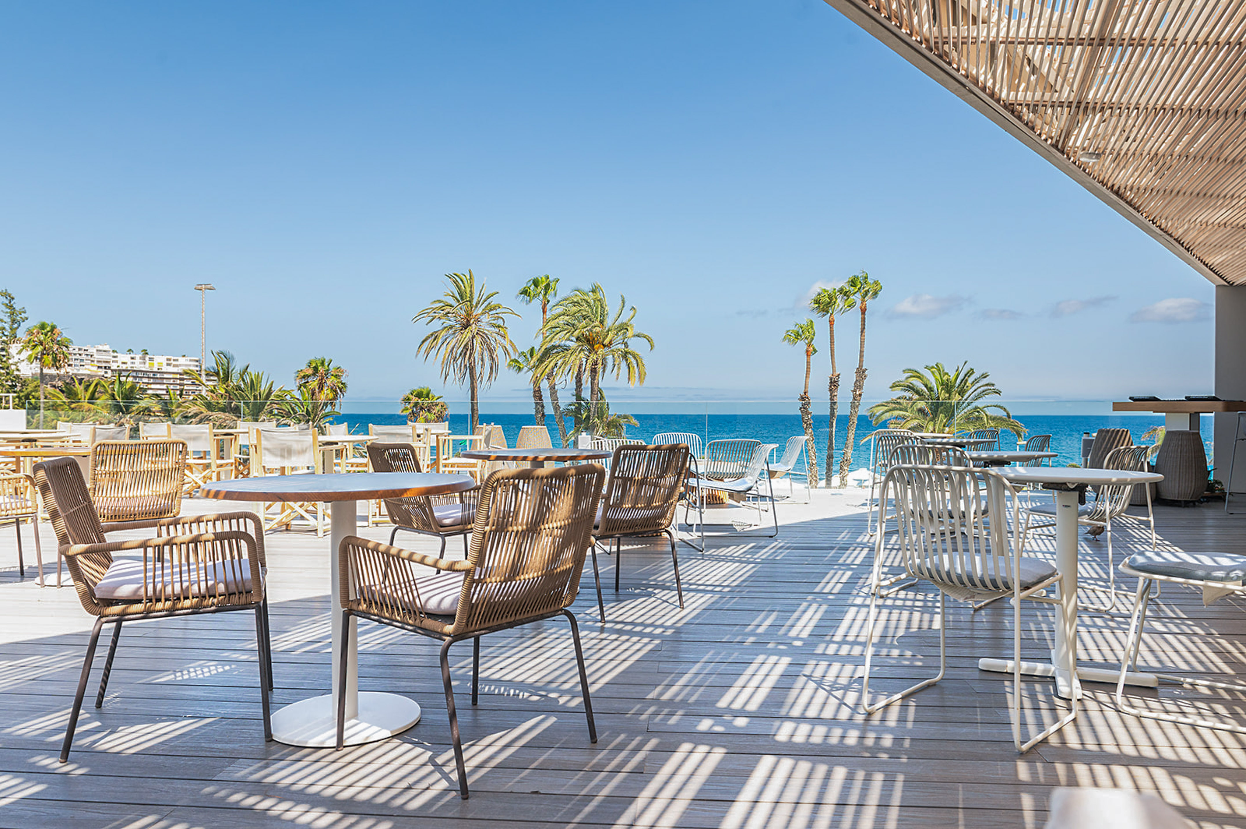 Outdoor restaurant terrace with ocean view, palm trees, and modern chairs.