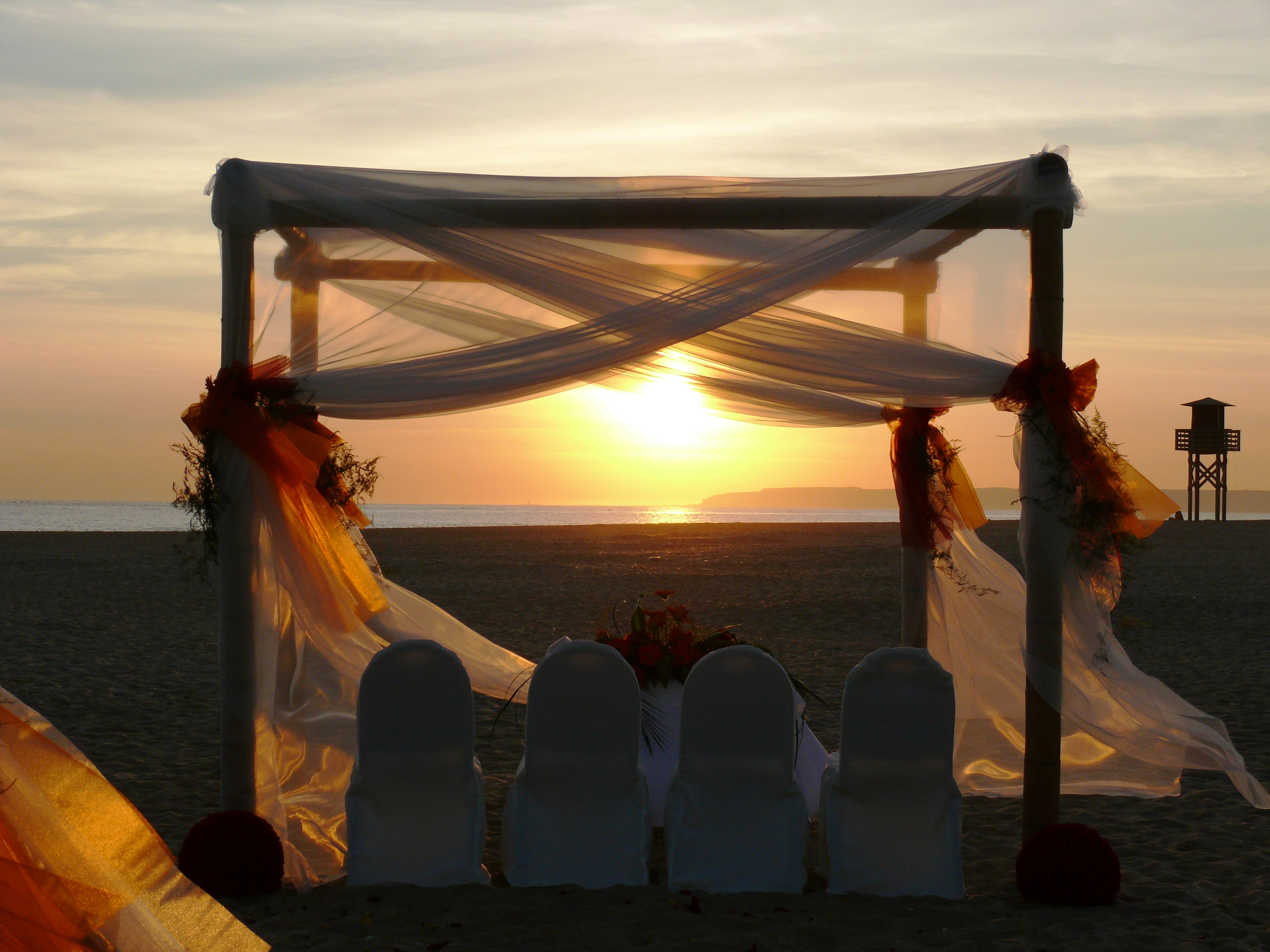 Sunset beach wedding arch with white drapes and chairs overlooking ocean.