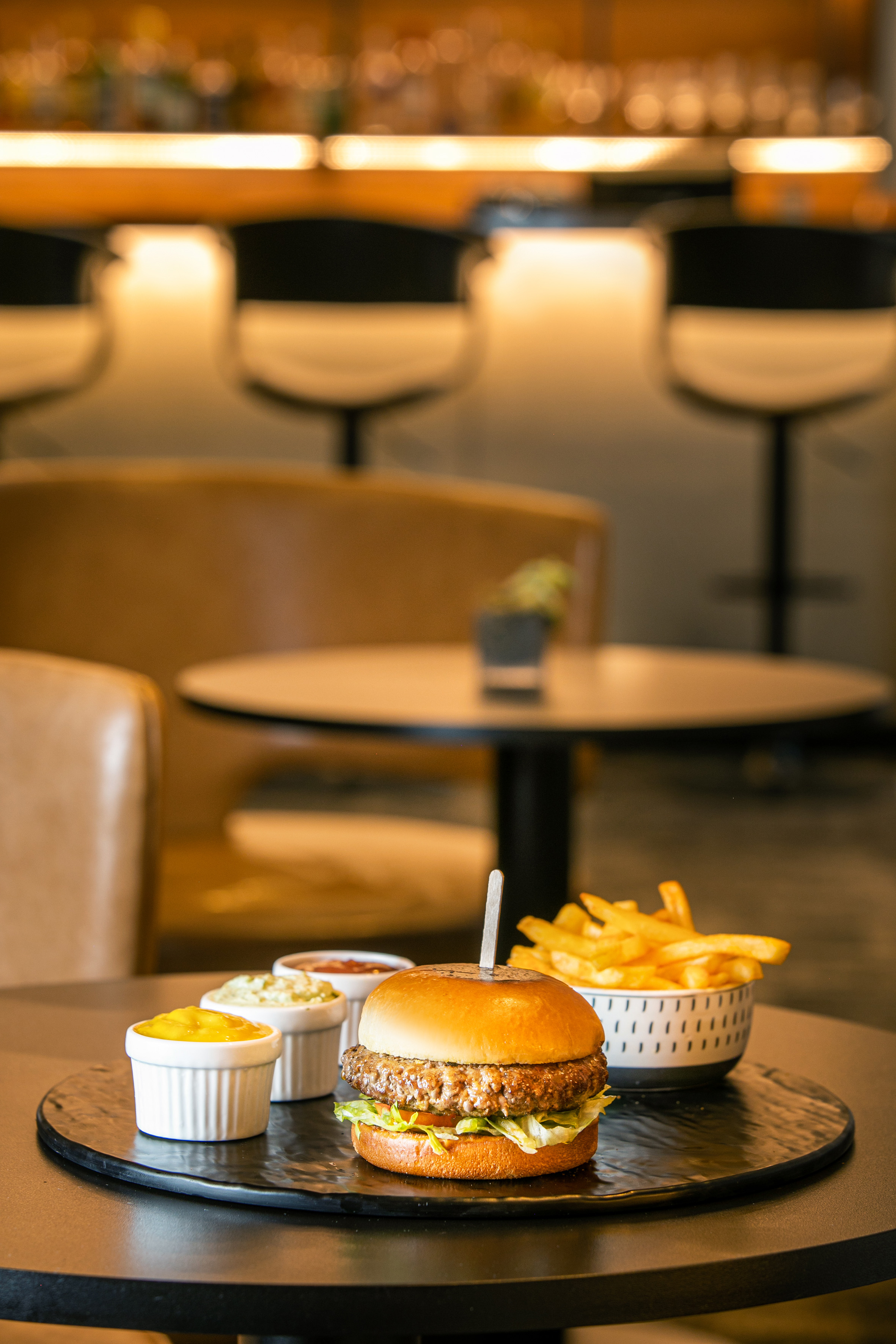 Burger, french fries, three sauces on black plate at restaurant table.