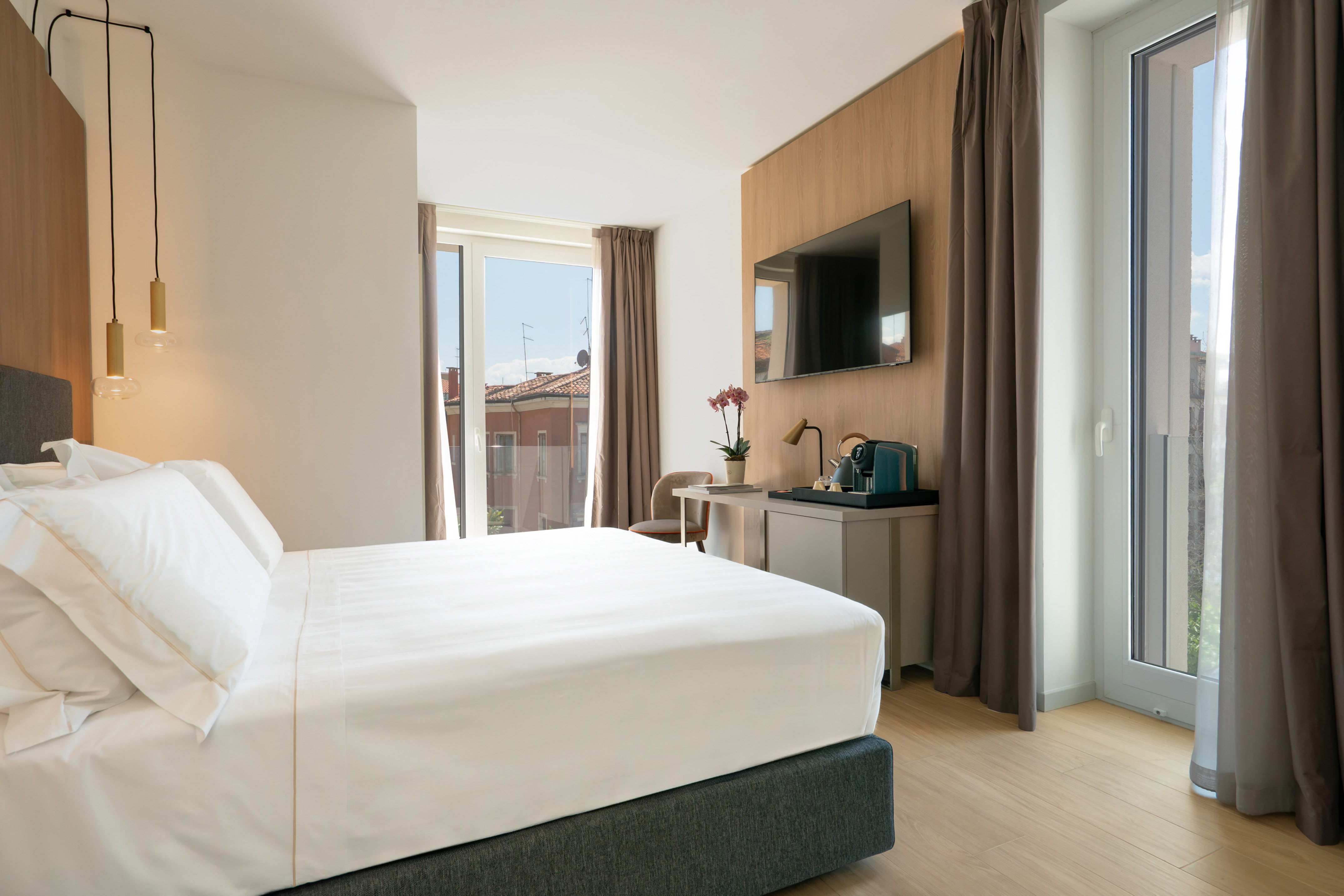 Modern hotel room with white bed, city view from window, TV, and coffee station.