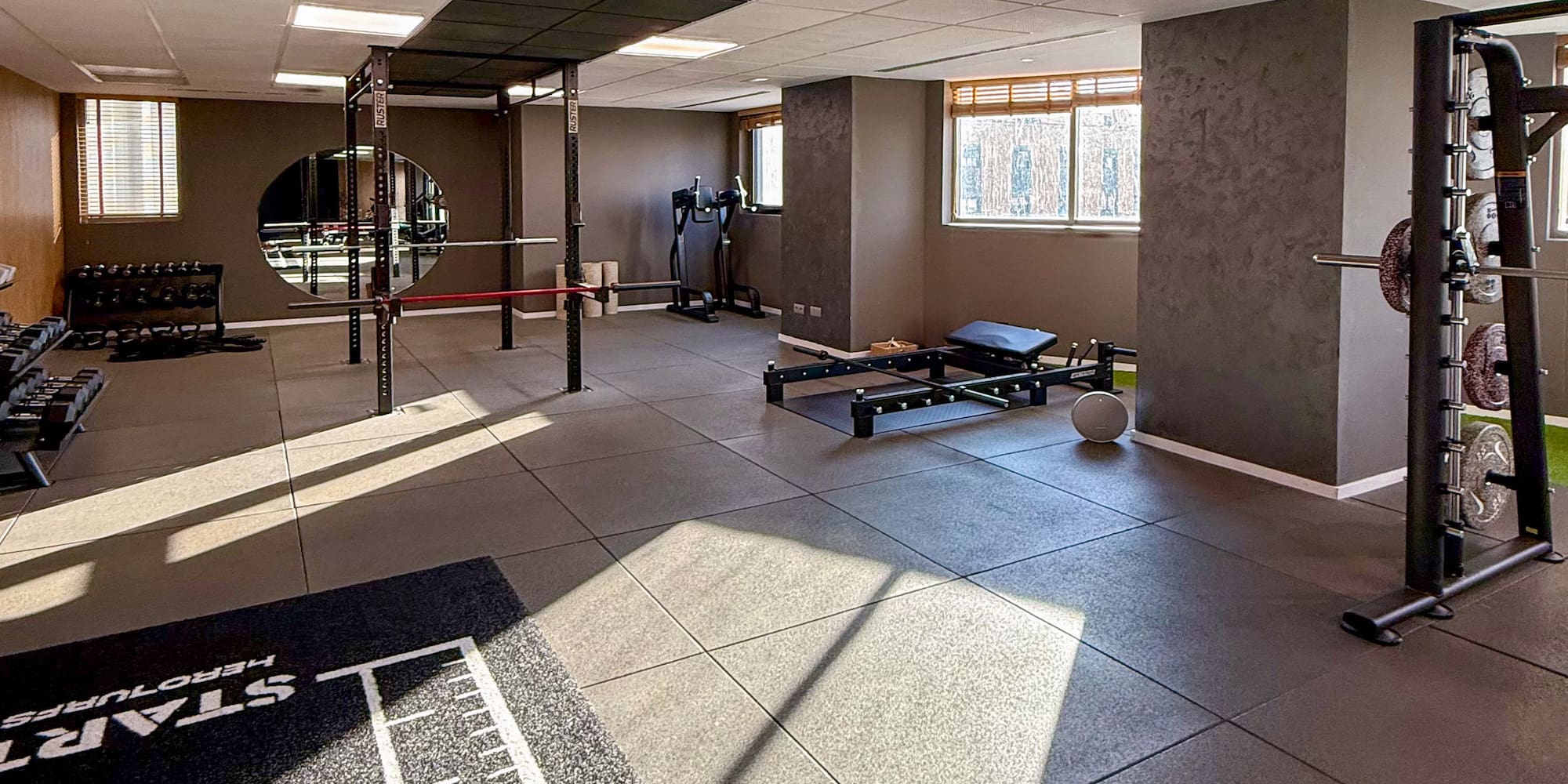 Modern gym interior with workout equipment, rubber floor, and sunny windows.
