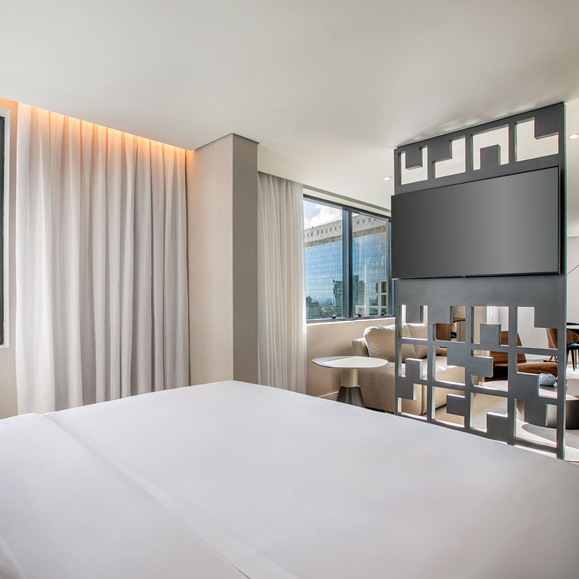 Modern hotel suite: bedroom with city view, TV, and adjoining living area.