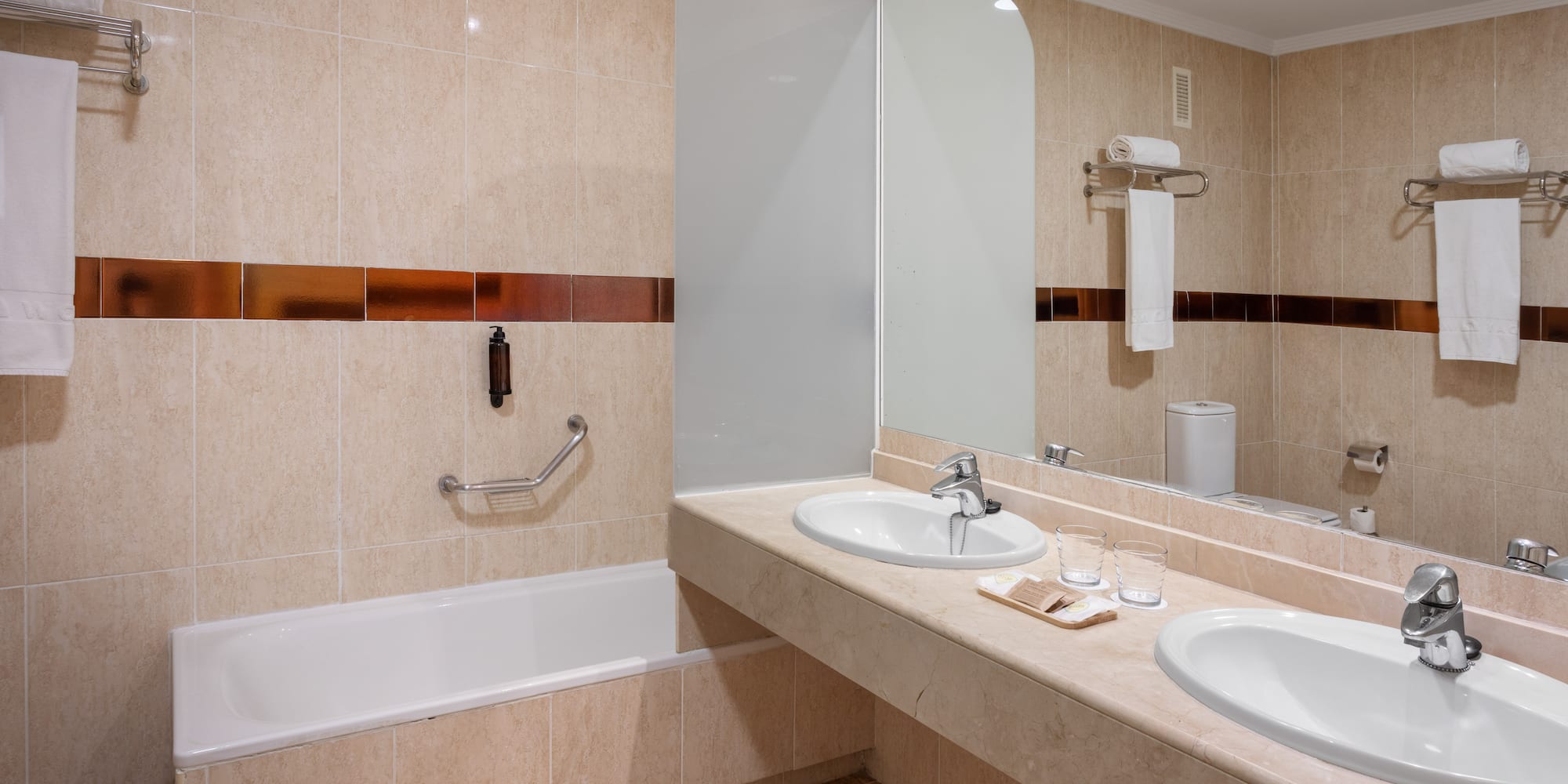 Modern hotel bathroom with double vanity, large mirror, bathtub, and beige tiled walls.