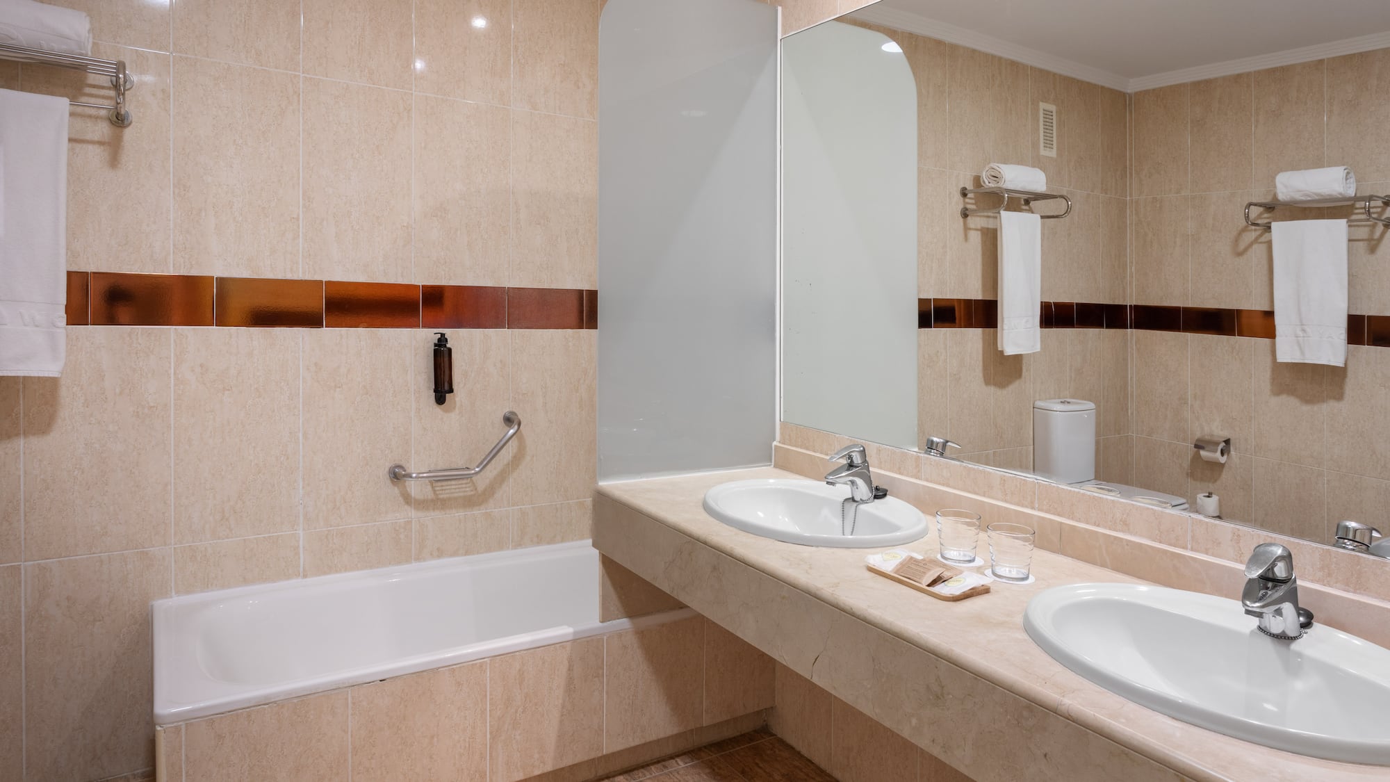 Modern hotel bathroom with double vanity, large mirror, bathtub, and beige tiled walls.
