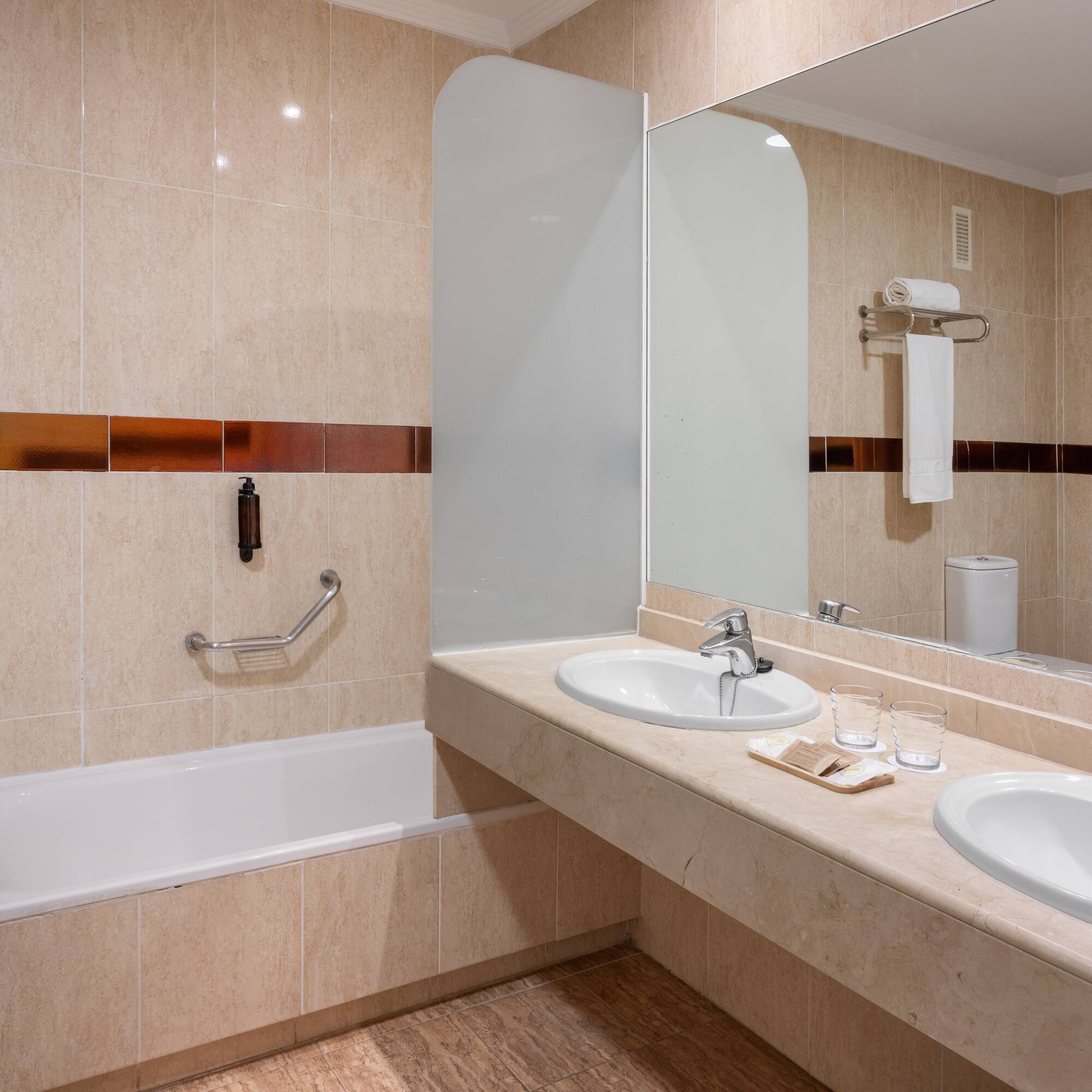 Modern hotel bathroom with double vanity, large mirror, bathtub, and beige tiled walls.