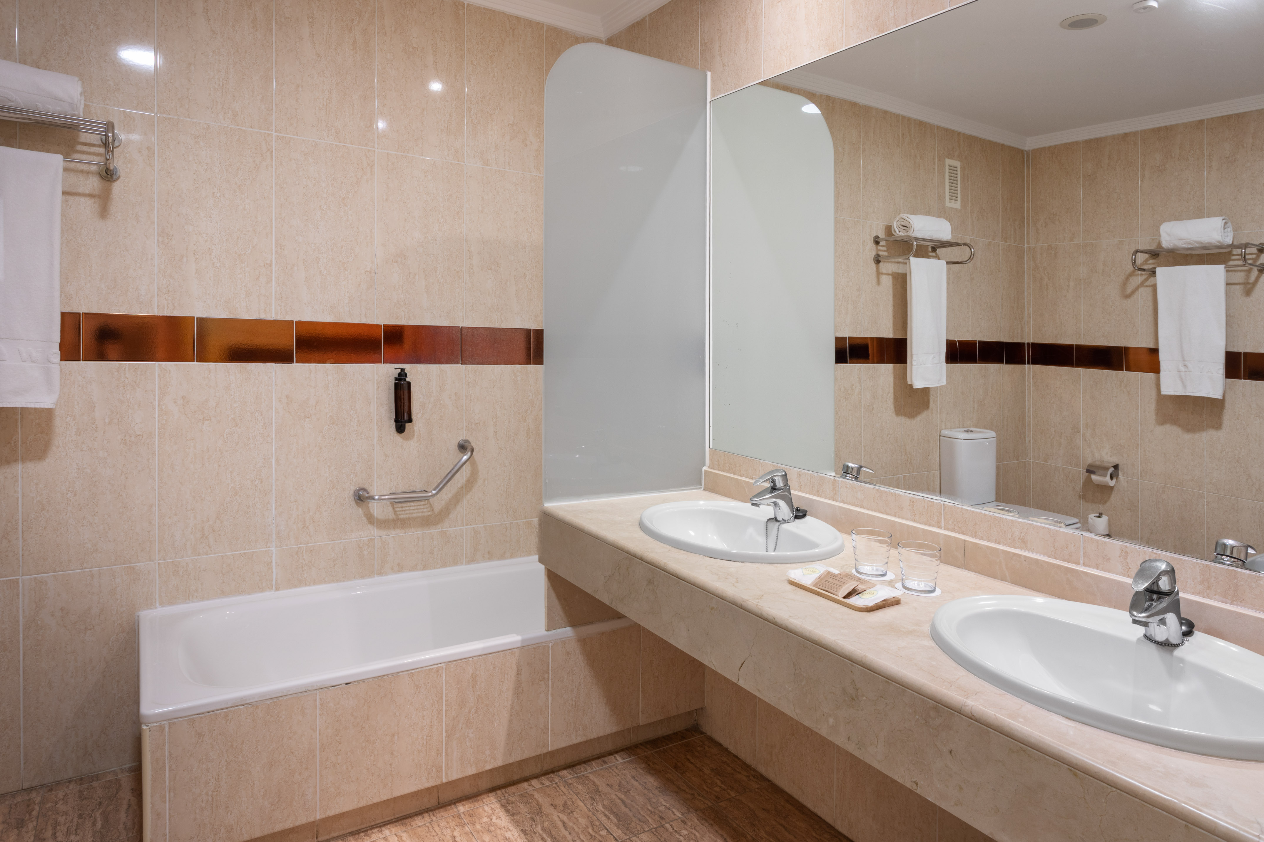 Modern hotel bathroom with double vanity, large mirror, bathtub, and beige tiled walls.