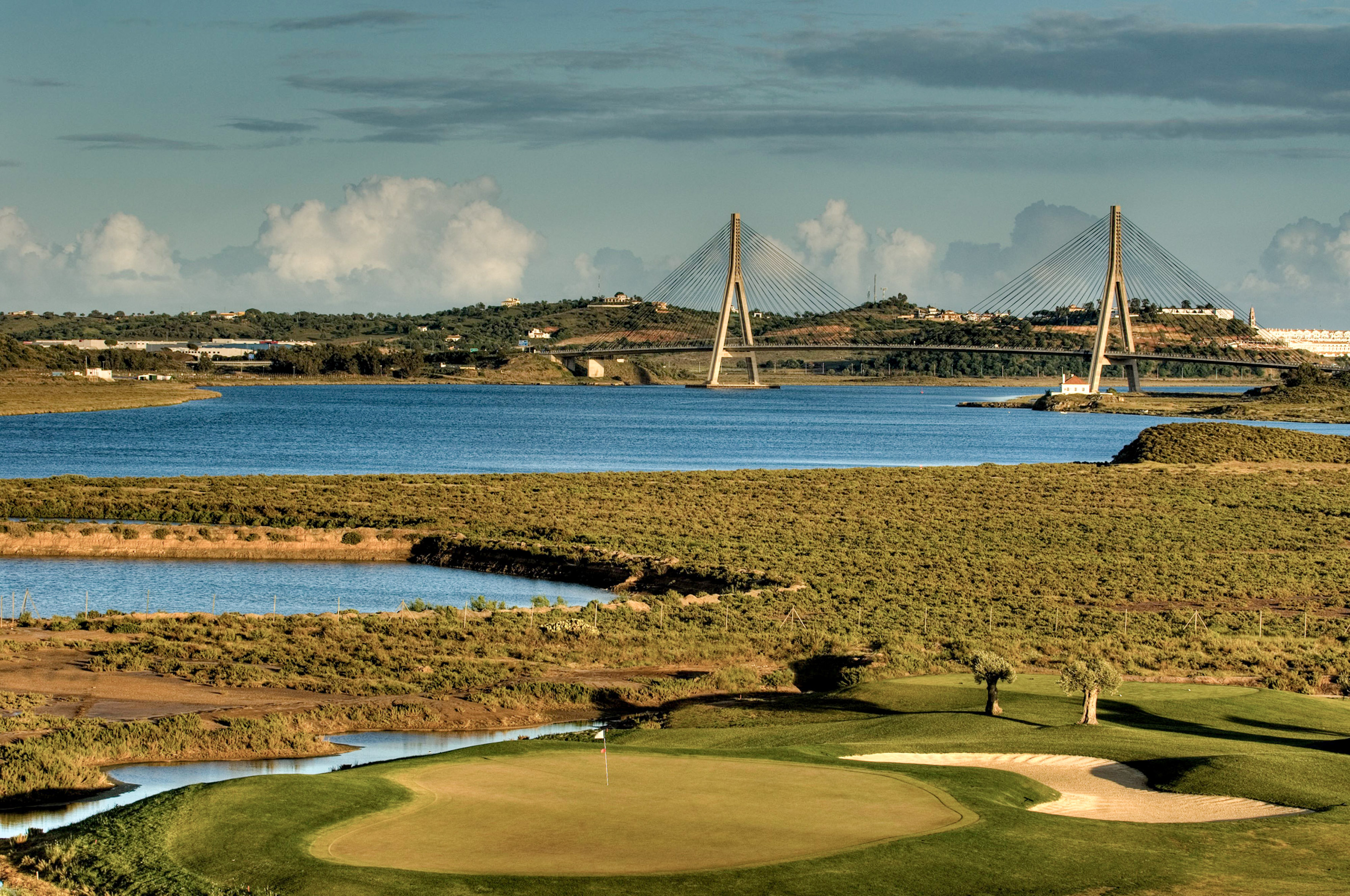 Golf course green, river, and cable-stayed bridge under cloudy sky.