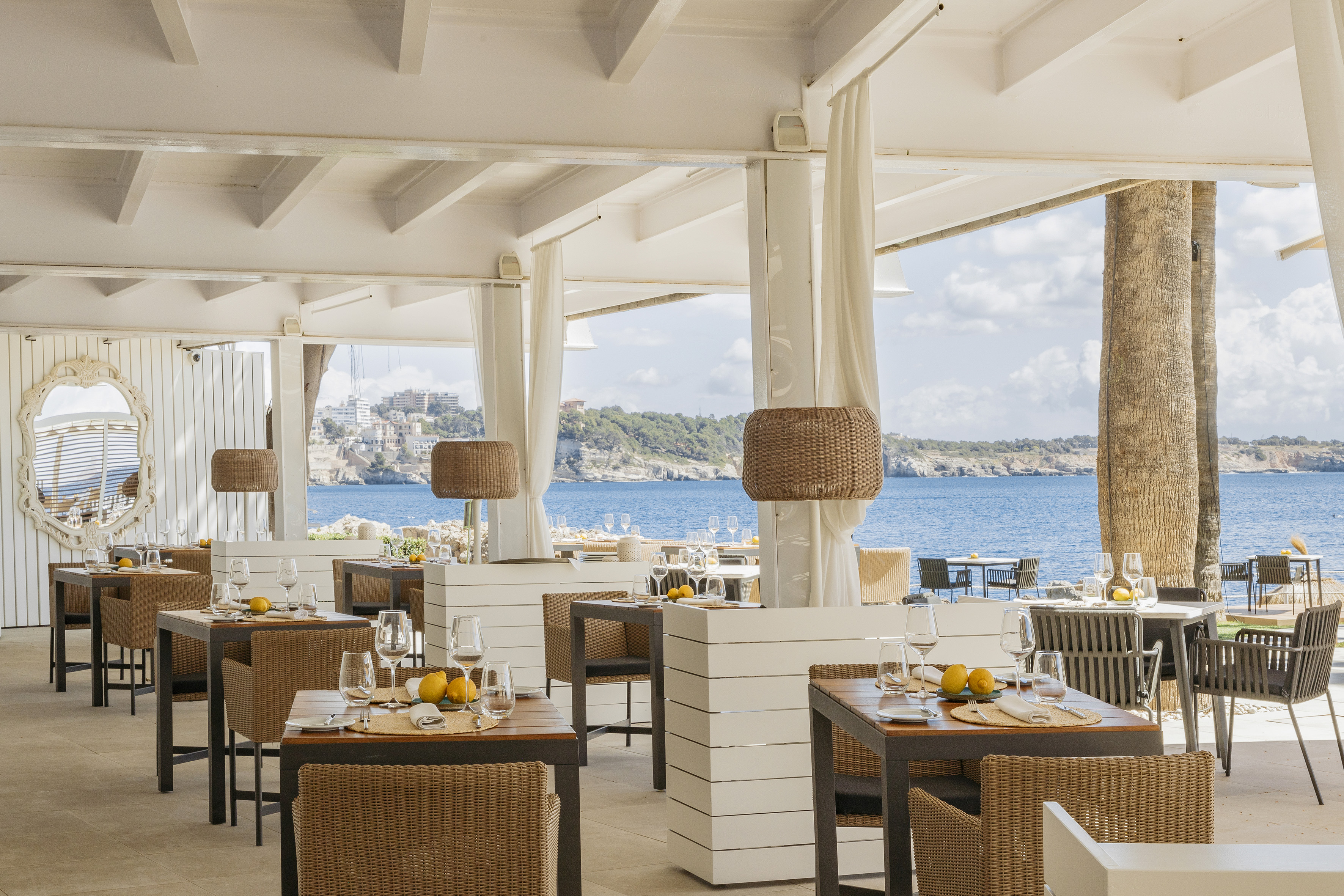 Outdoor restaurant terrace with sea view, set tables, wicker chairs, white decor.