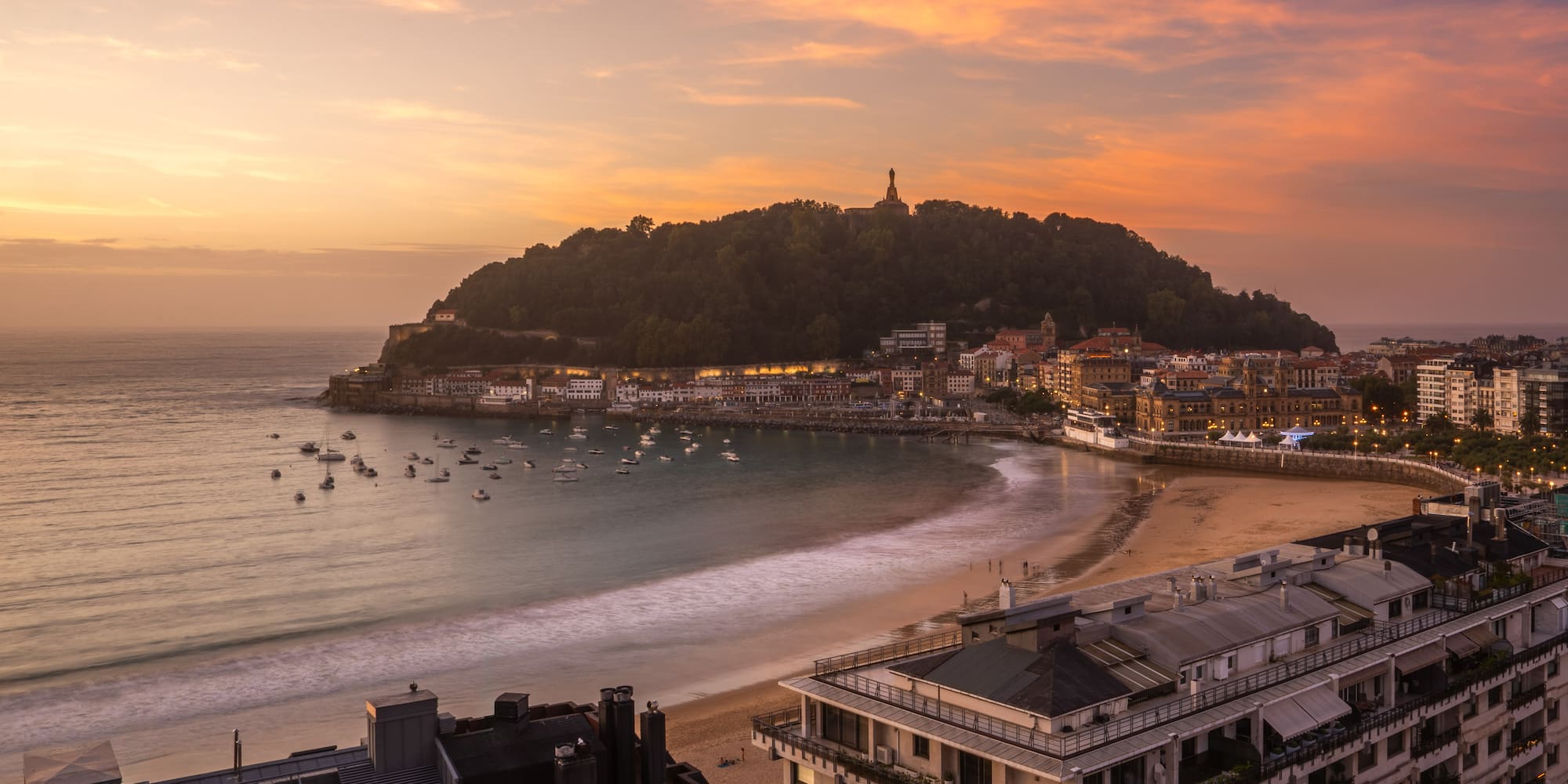 Vista aérea atardecer Bahía La Concha, San Sebastián, barcos y ciudad iluminada.