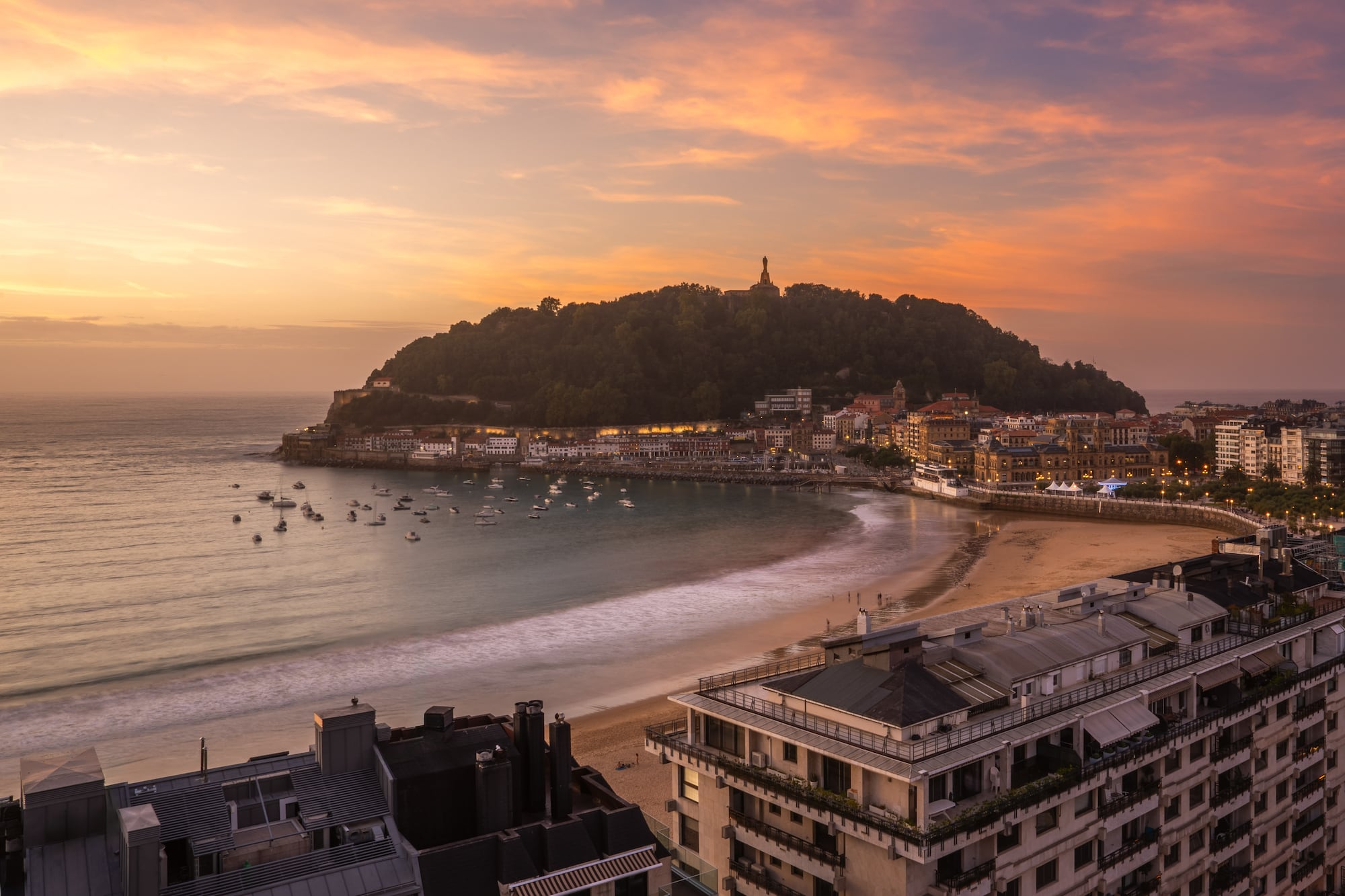 Vista aérea atardecer Bahía La Concha, San Sebastián, barcos y ciudad iluminada.