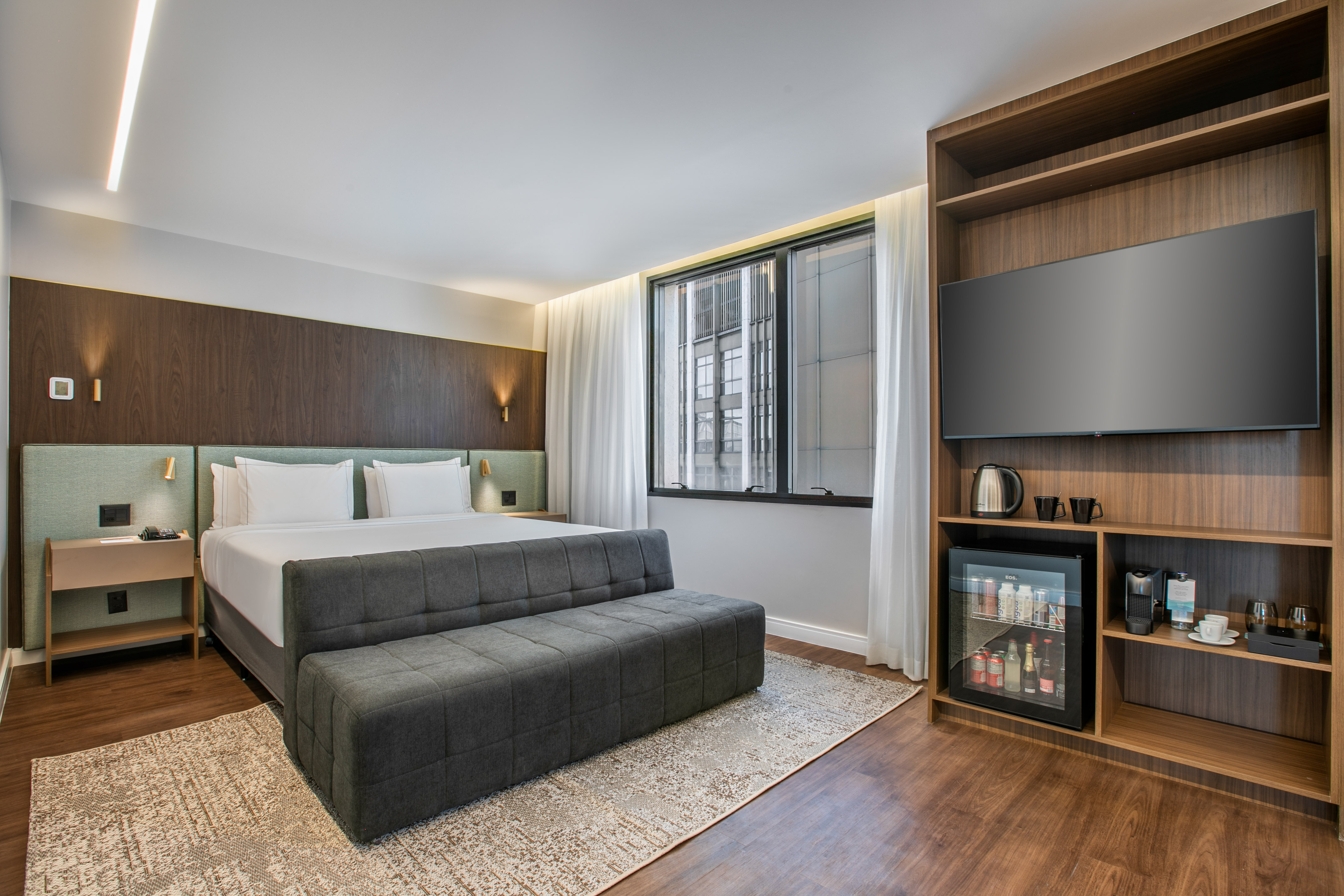Modern hotel room: king bed, TV, mini-fridge, coffee station, wood paneling.
