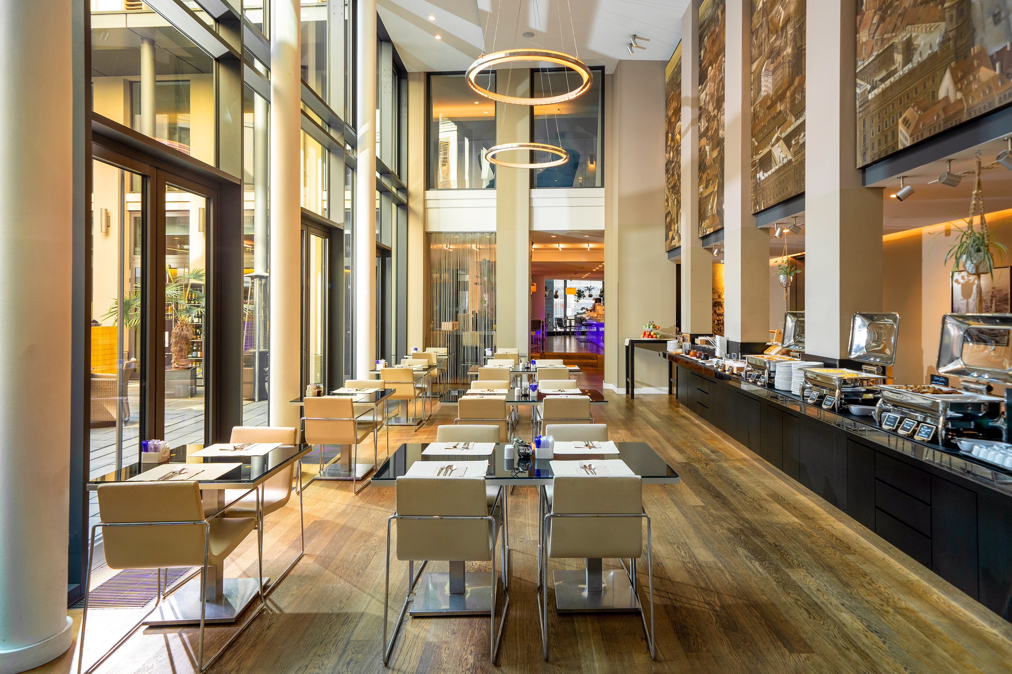 Modern hotel restaurant with a buffet line, set dining tables, and large windows.