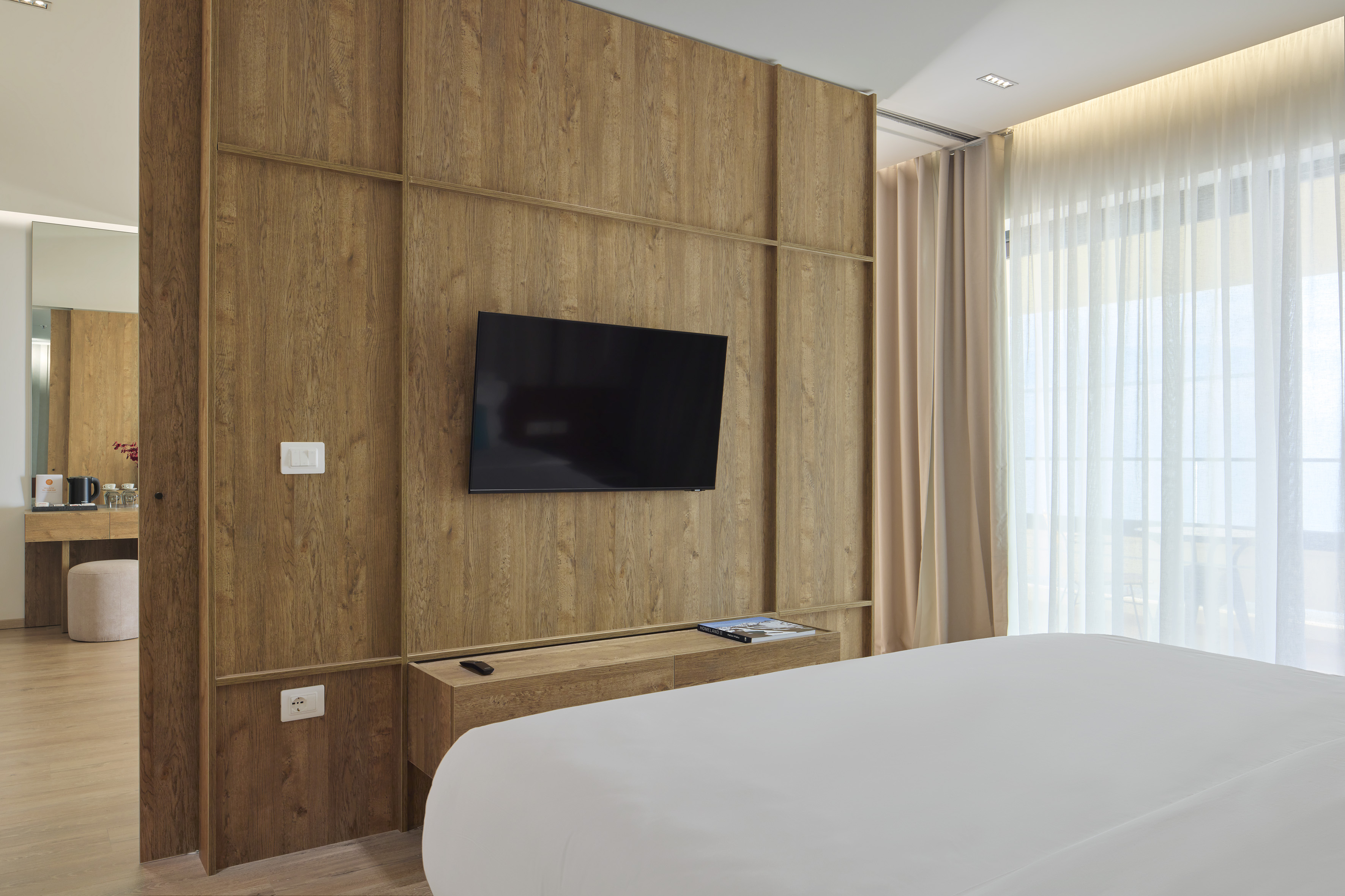 Modern hotel bedroom with wood paneling, mounted TV, and white bed.