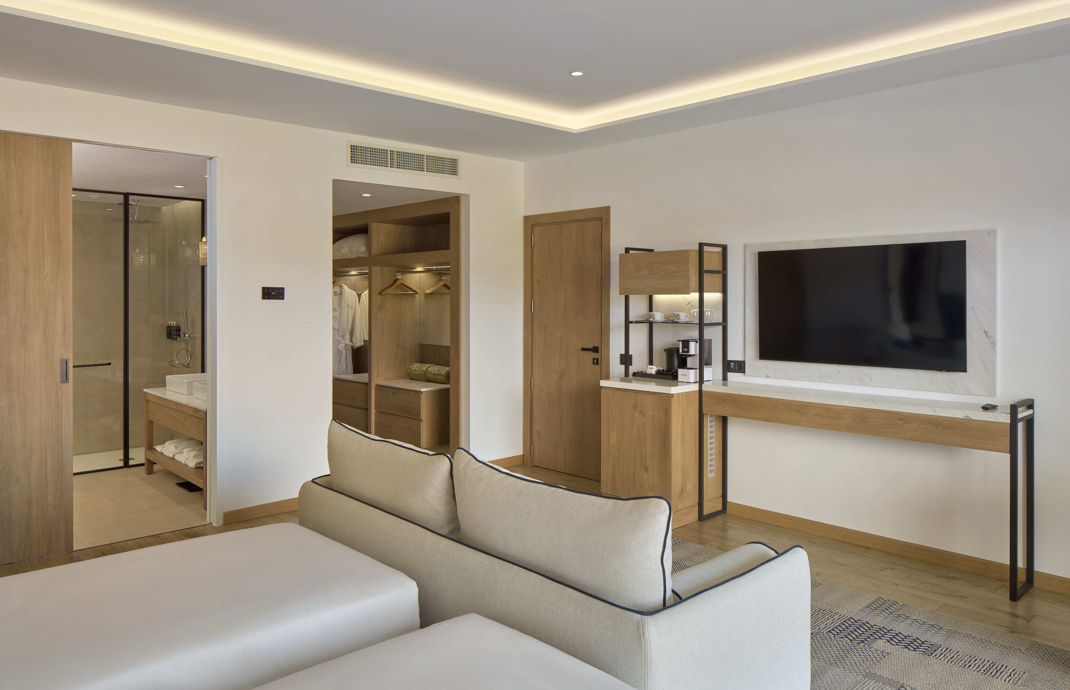 Modern hotel room with sofa, wall TV, coffee station, open wardrobe, and bathroom access.