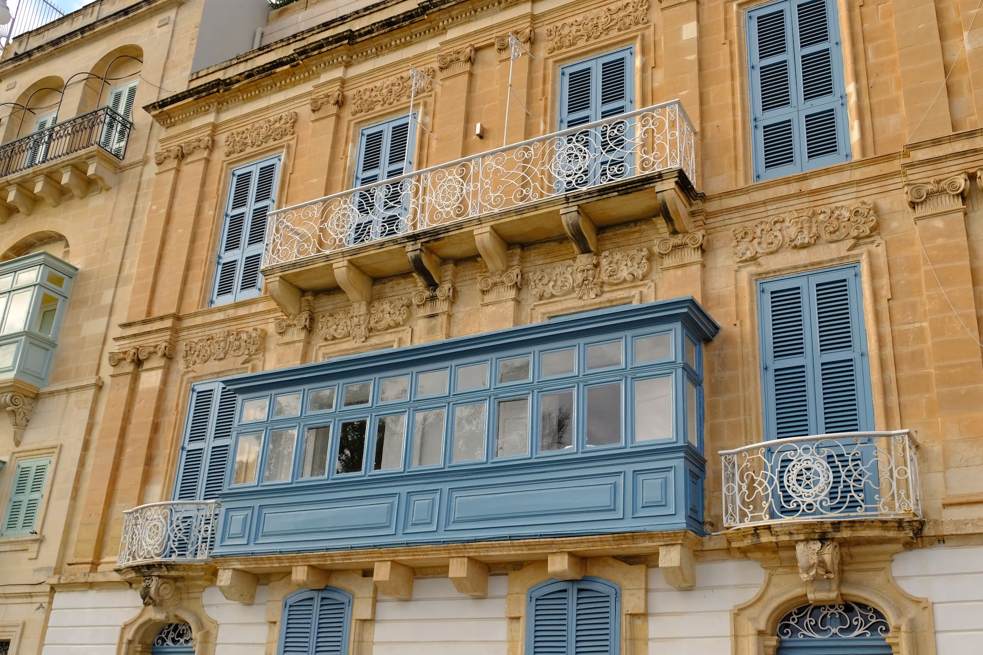 Edificio maltés de piedra con balcones tradicionales y persianas azules.