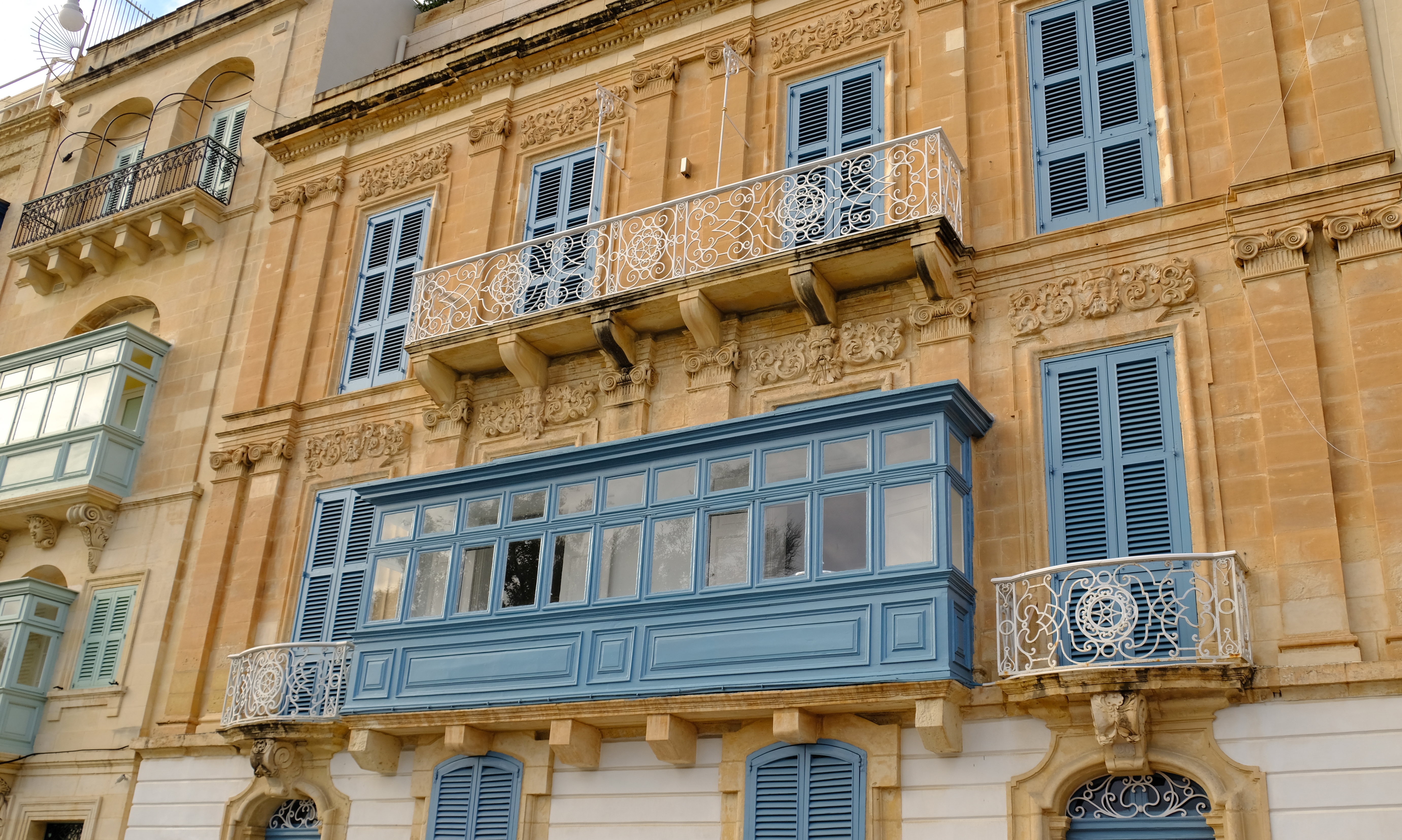 Edificio maltés de piedra con balcones tradicionales y persianas azules.