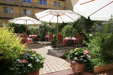 Shaded outdoor patio with white umbrellas, dining tables, chairs, and vibrant plants.