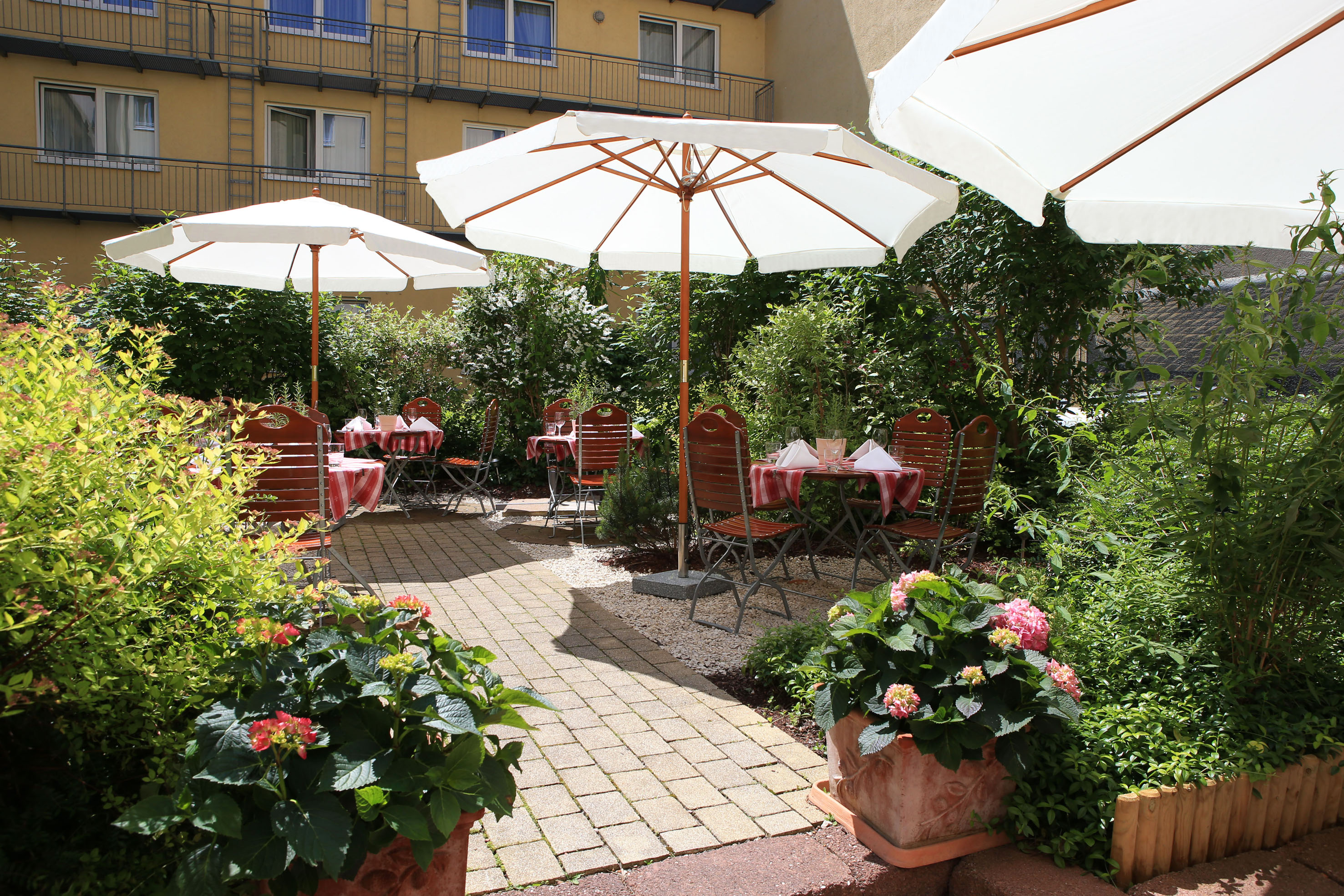 Shaded outdoor patio with white umbrellas, dining tables, chairs, and vibrant plants.