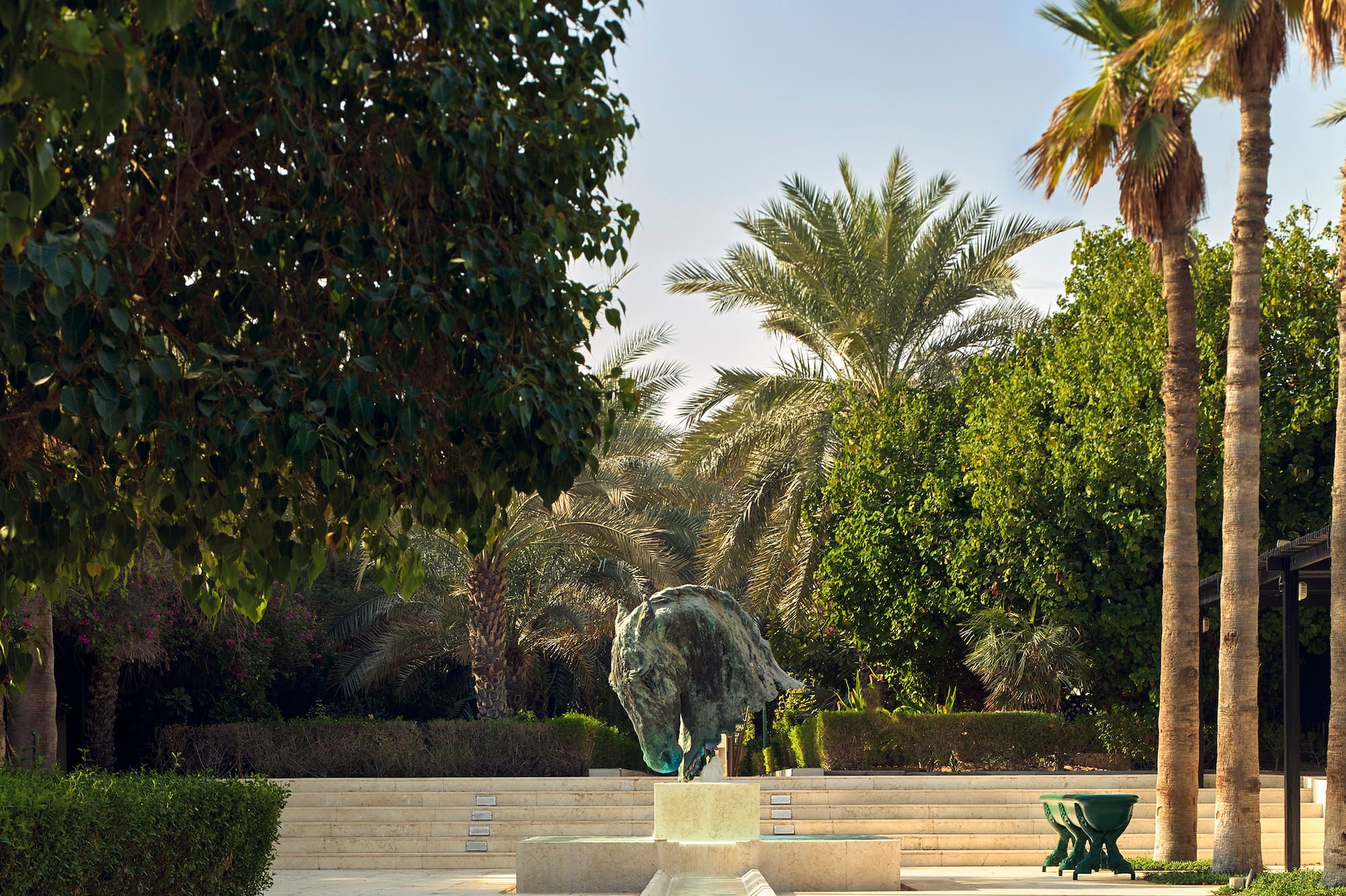 Bronze horse head sculpture fountain in lush garden with water channel, palm trees.