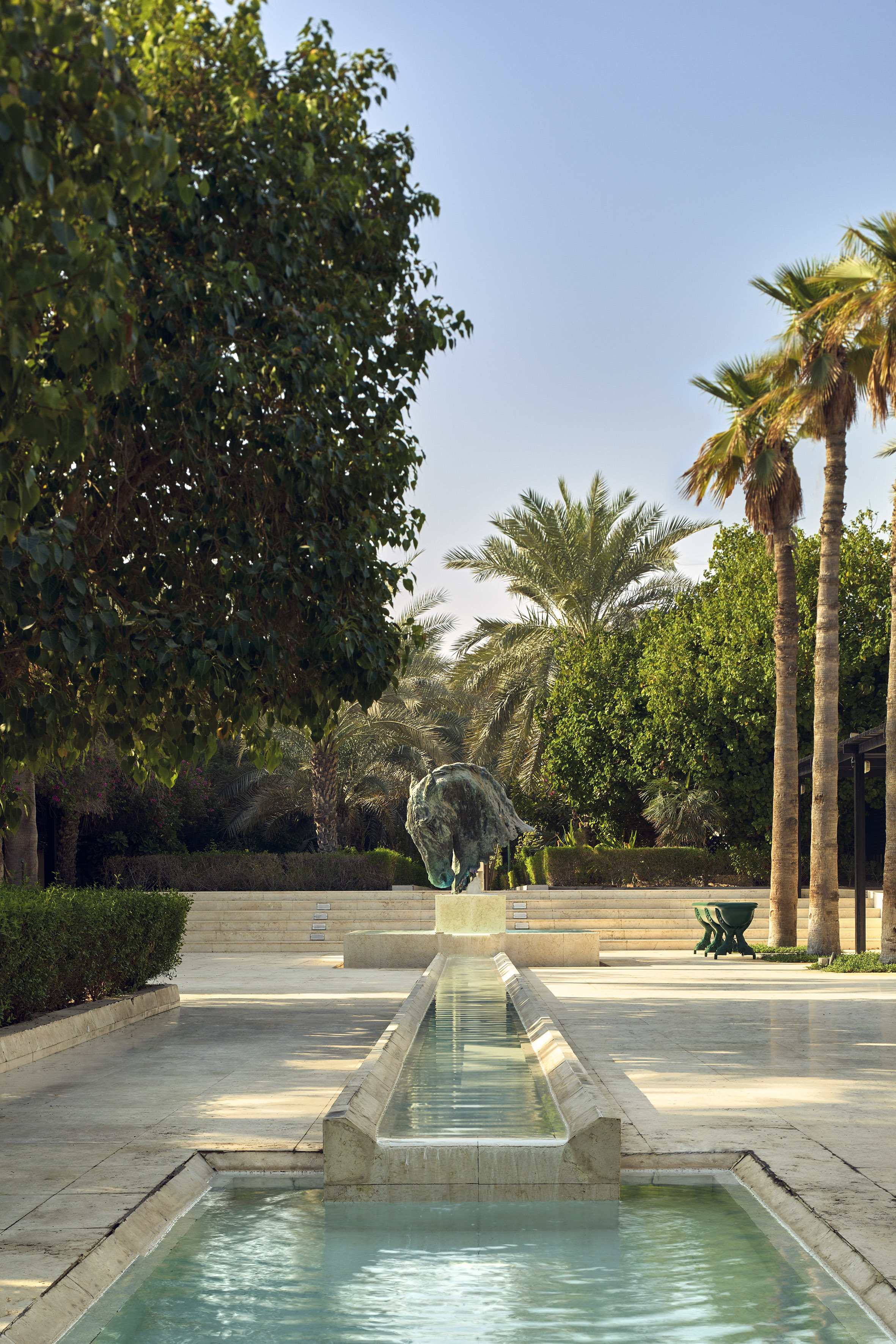 Bronze horse head sculpture fountain in lush garden with water channel, palm trees.